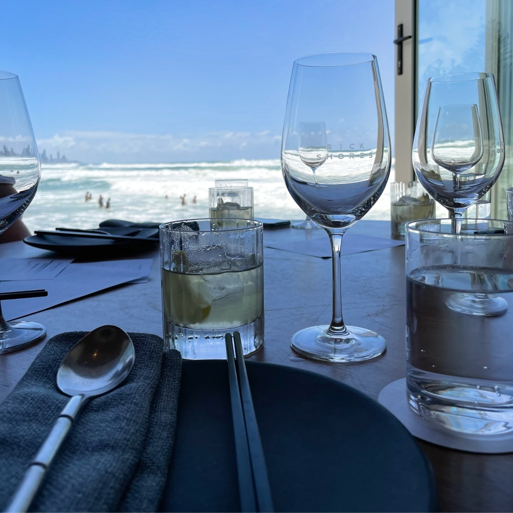 A table looking out to the beach, people in the surf further away and a clear blue sky. A black plate is in the foreground with chopsticks, large spoon and napkin on it. A tumbler with a yellow liquid is in front as are two empty wine glasses. Within the wine glasses are the smaller images of other wine glasses that are sitting behind them.