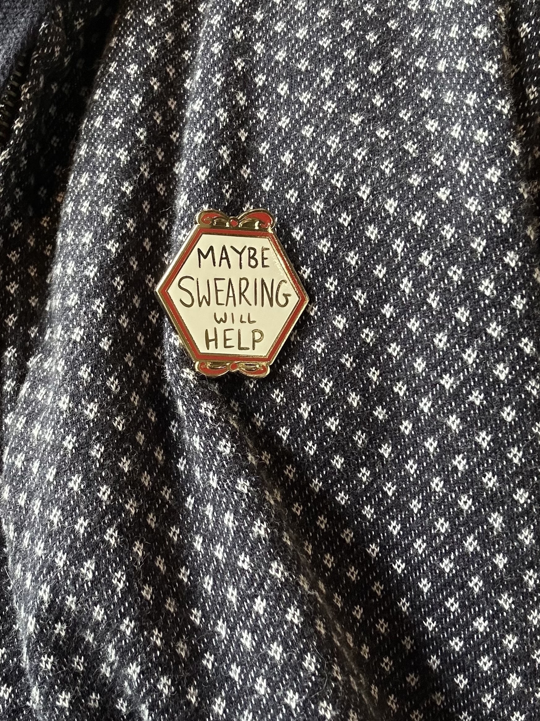 A hexagon shaped badge pinned on a black and white jumper. The badge is white with a red border and has “maybe swearing will help” in gold letters.