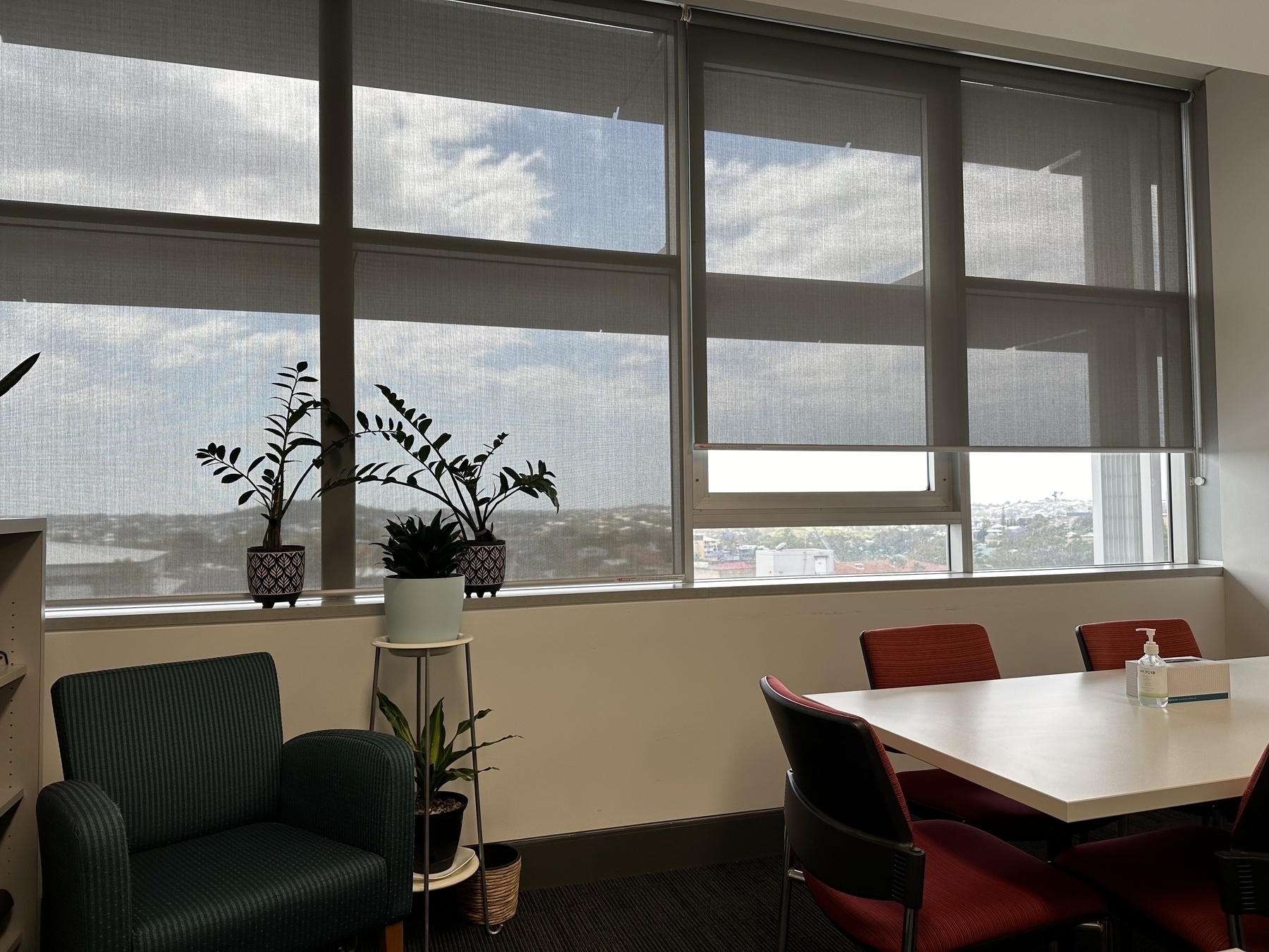Looking across an office towards windows with roller blinds partially down. A white meeting table with red chairs to the right. A green chair with plants near it on the left. 