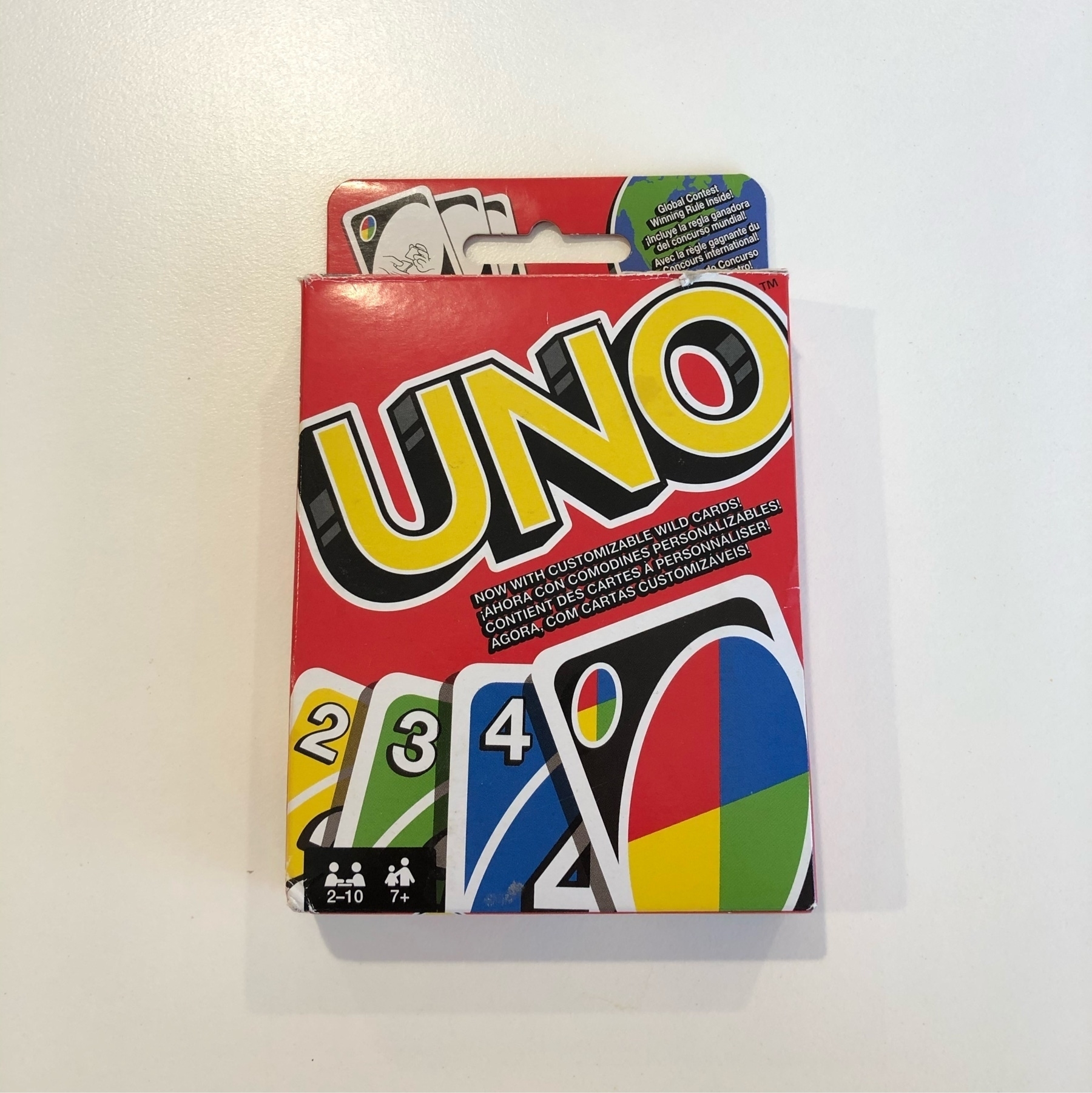A box of Uno playing cards on a white table. The box is red with UNO in large yellow letters in the top half and the bottom has yellow, green and blue cards numbered 2, 3 and 4.