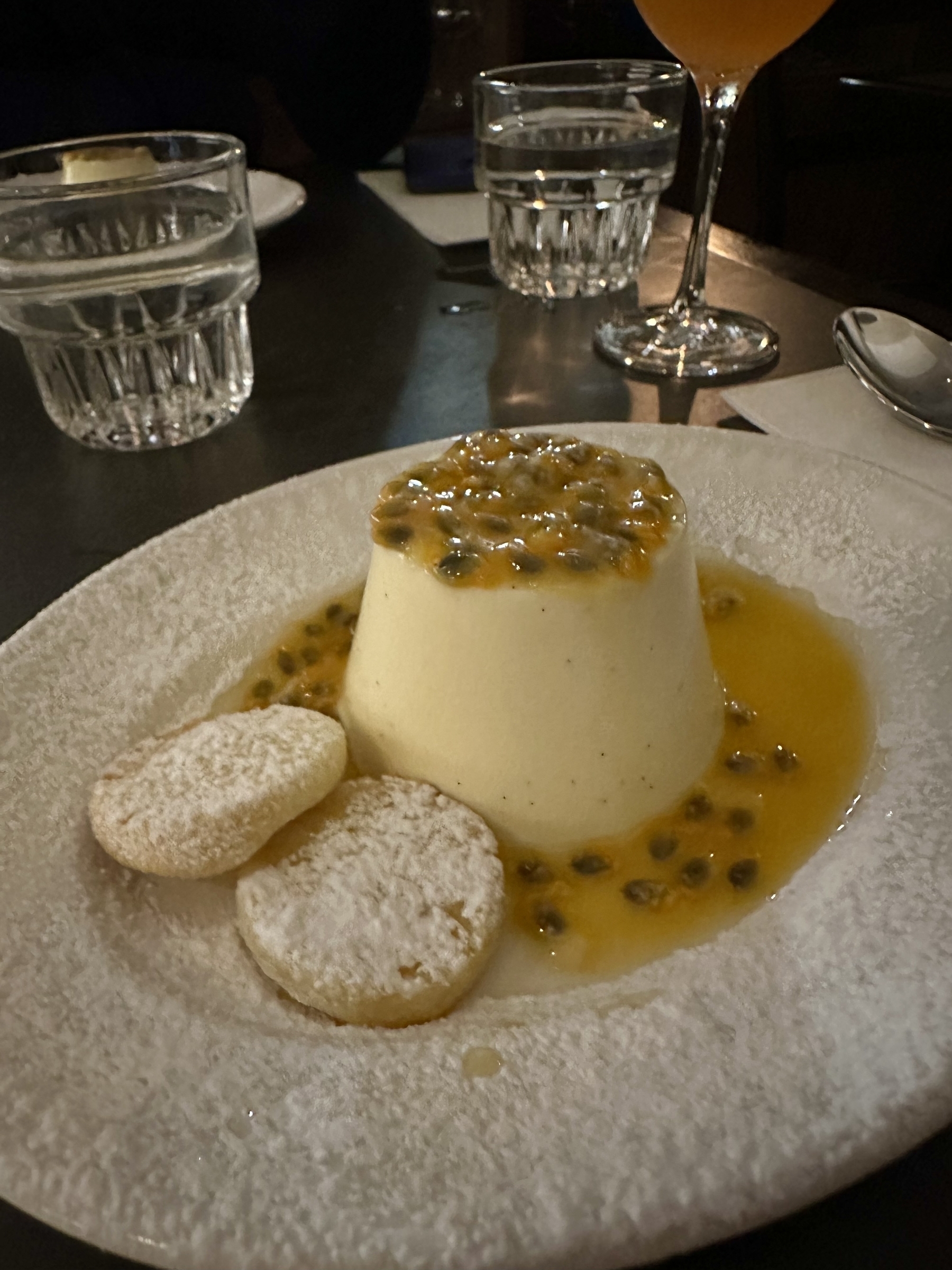 A vanilla white coloured soft almost cylindrical dessert sitting in a shallow plate with passion fruit pulp surrounding it and layered on top. Two small round shortbread biscuits are beside it and the plate is lightly dusted in flour.