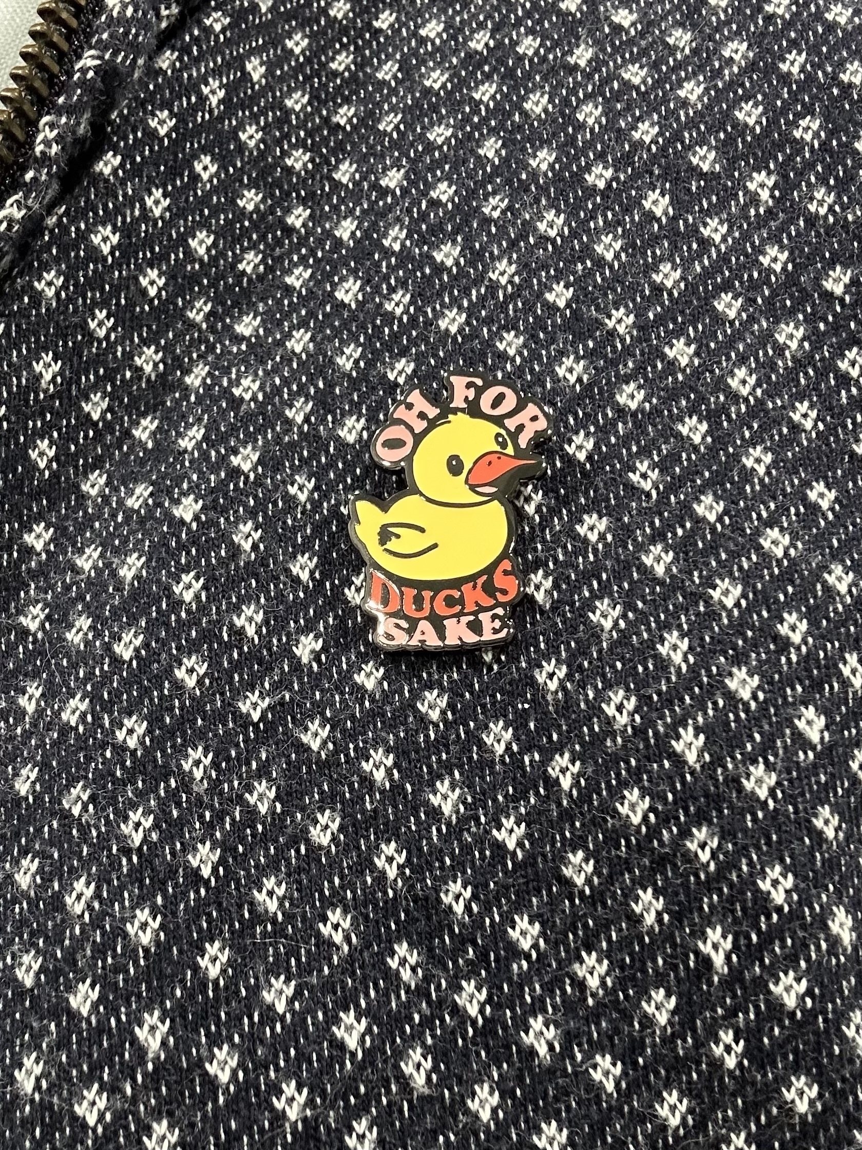 An enamel badge in the shape of a yellow duck with “Oh for ducks sake” around it.