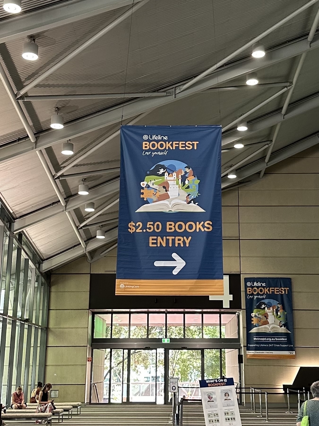 A large blue banner hanging from the ceiling of a convention centre. The banner has “Lifeline Bookfest. $2.50 books entry” and an arrow pointing right.