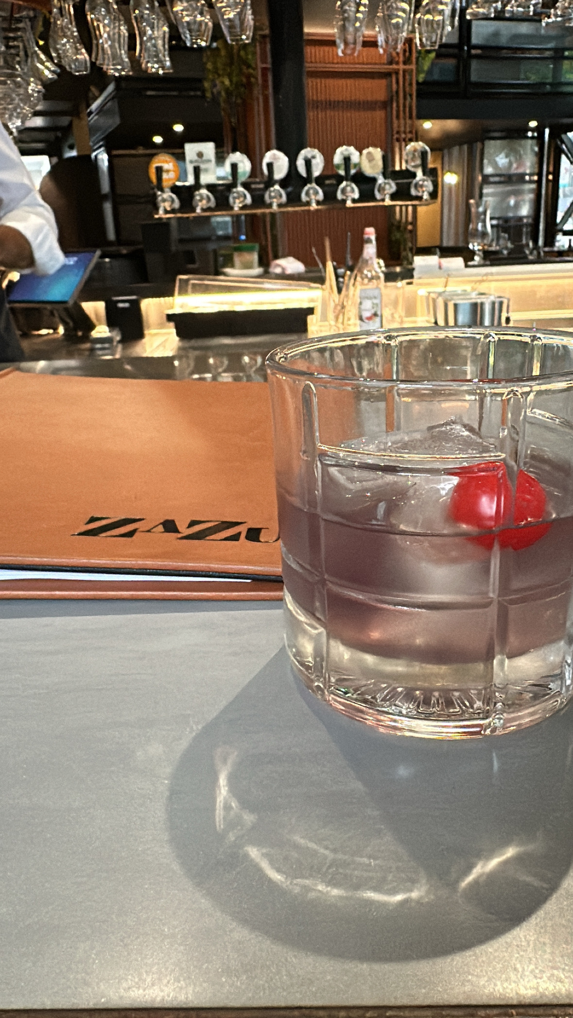 A bar counter with a cocktail with a red cherry in it. A tan covered menu behind it with the name of the bar - Zazu. In the background are beer taps on the other side of the bar.