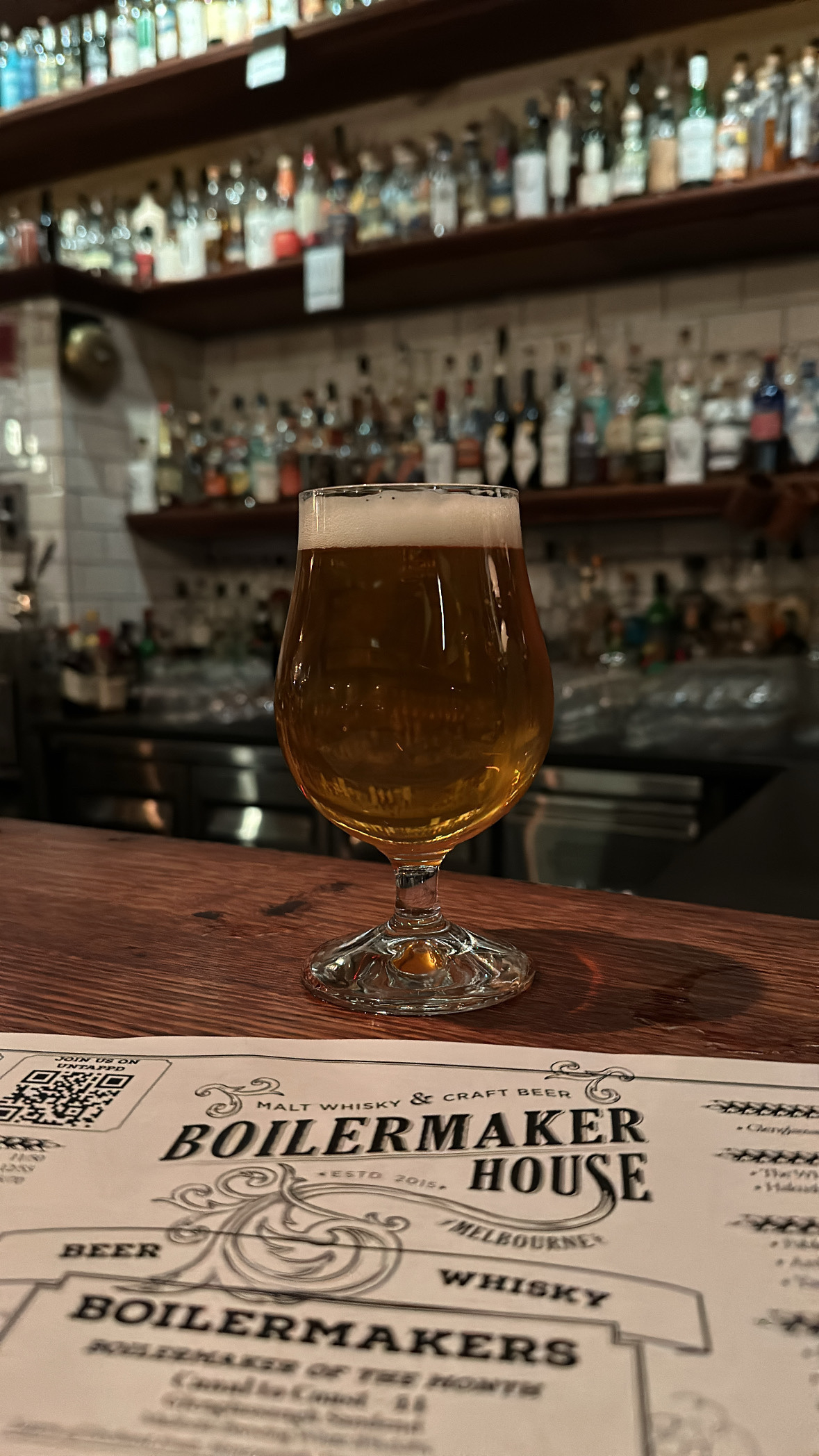 A beer glass sitting on a bar, shelves full of whisky bottles in the background. Part of the menu is in front of the glass displaying the name “Boilermaker House”.