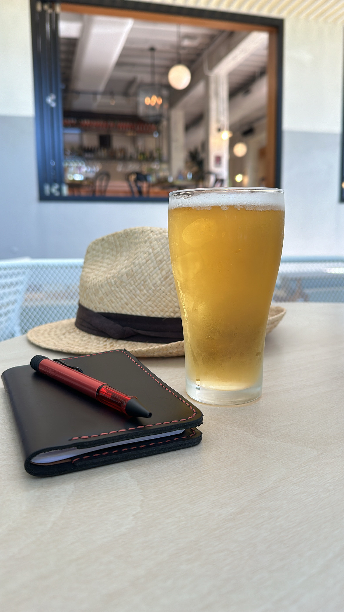 A straw hat, black A6 notebook with red pen on top, and a schooner of beer on a table. In the background is a large window with a bar visible inside.