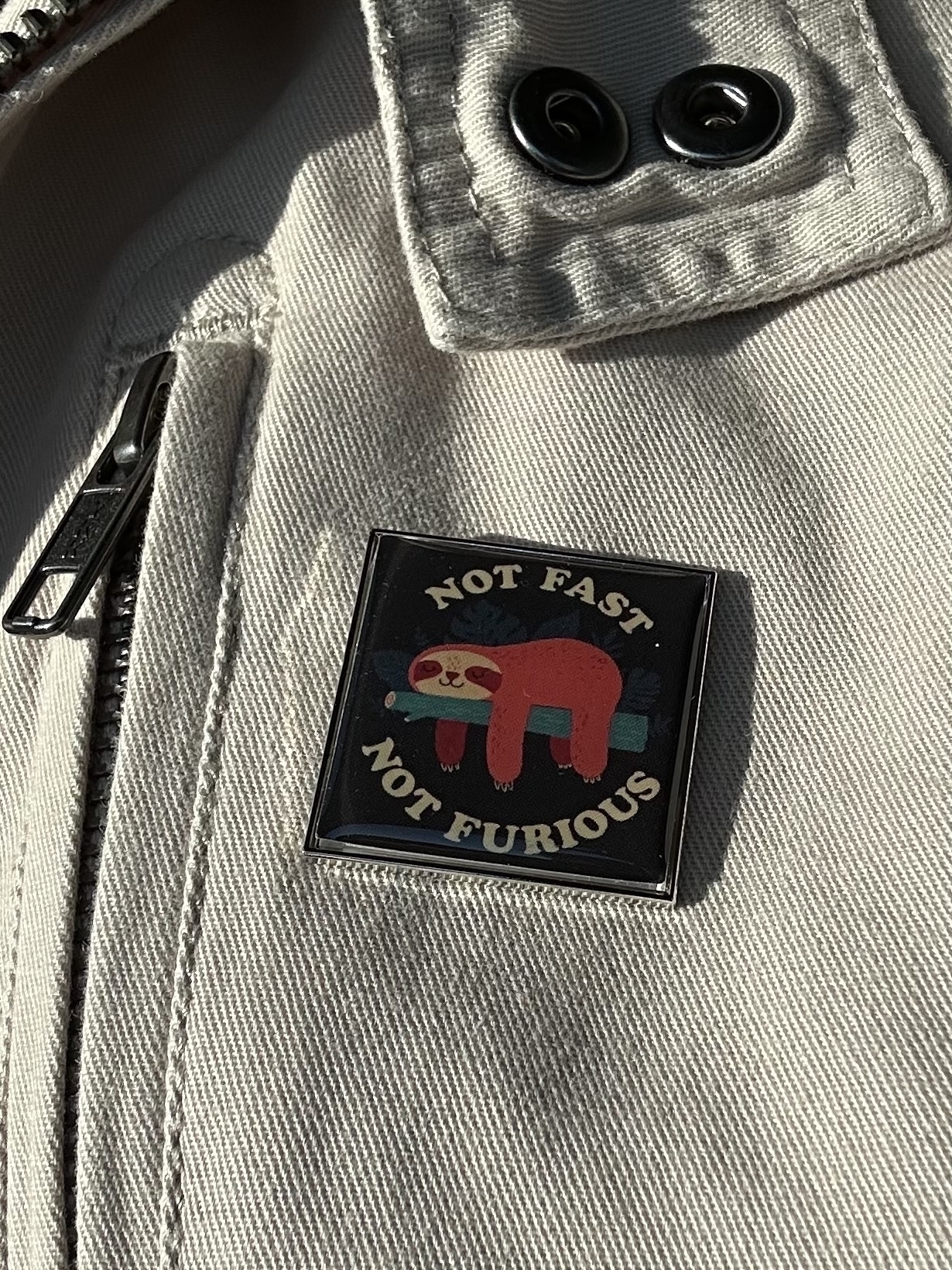 An enamel badge pinned on a cream jacket. The badge is black background, a red sloth draped on a green branch and “not fast” above and then “not furious” below.