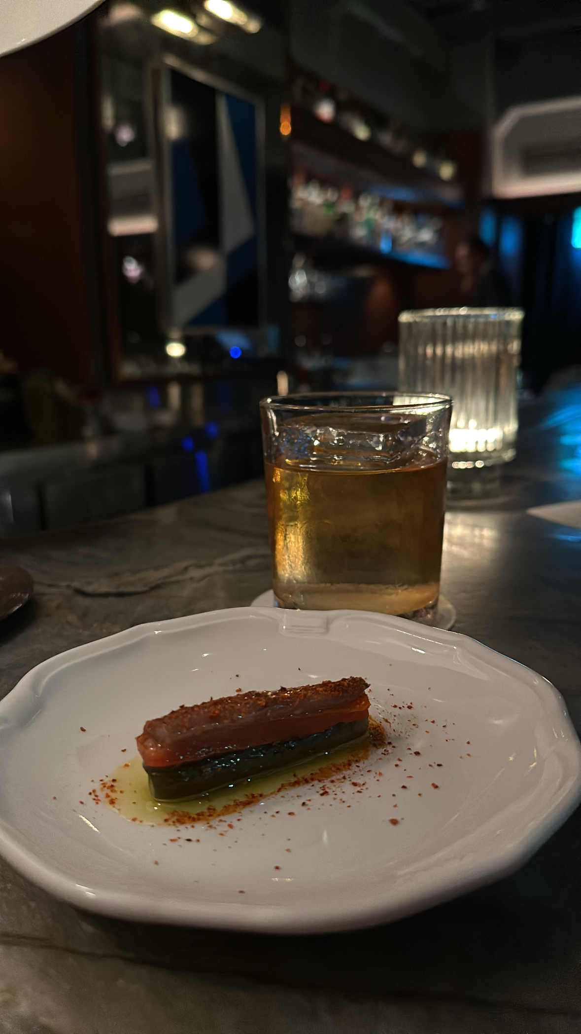 A marble top bar. In the foreground is a white plate with a bar snack and immediately behind it is a whisky drink with a large cube of ice. There’s a glass candlelight on the bar behind and in the background are the dimly lit bar shelves and bottles.