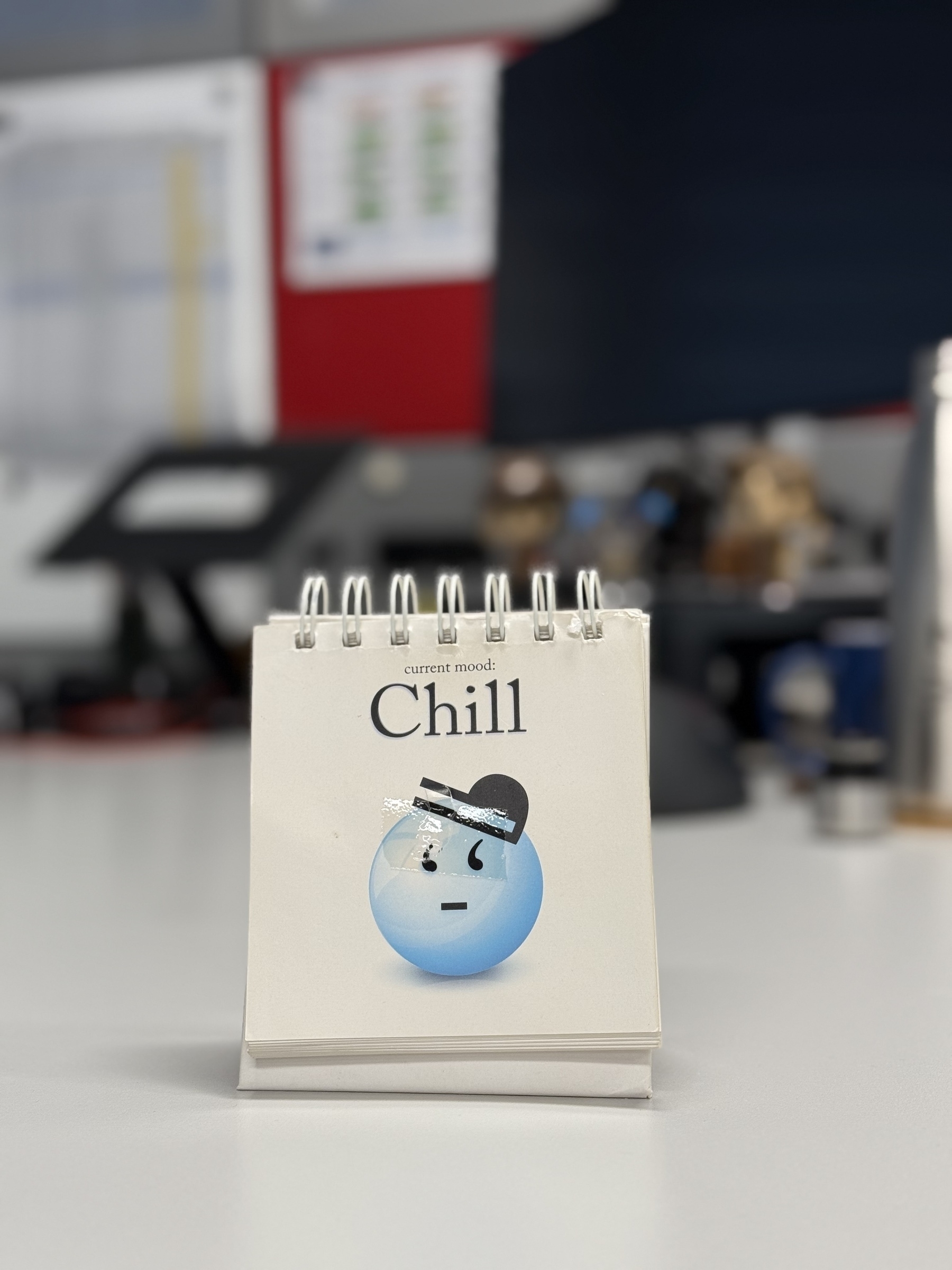 A sign sitting on an office desk. White background, blue emoji face and the words “Current Mood: CHILL” above it.