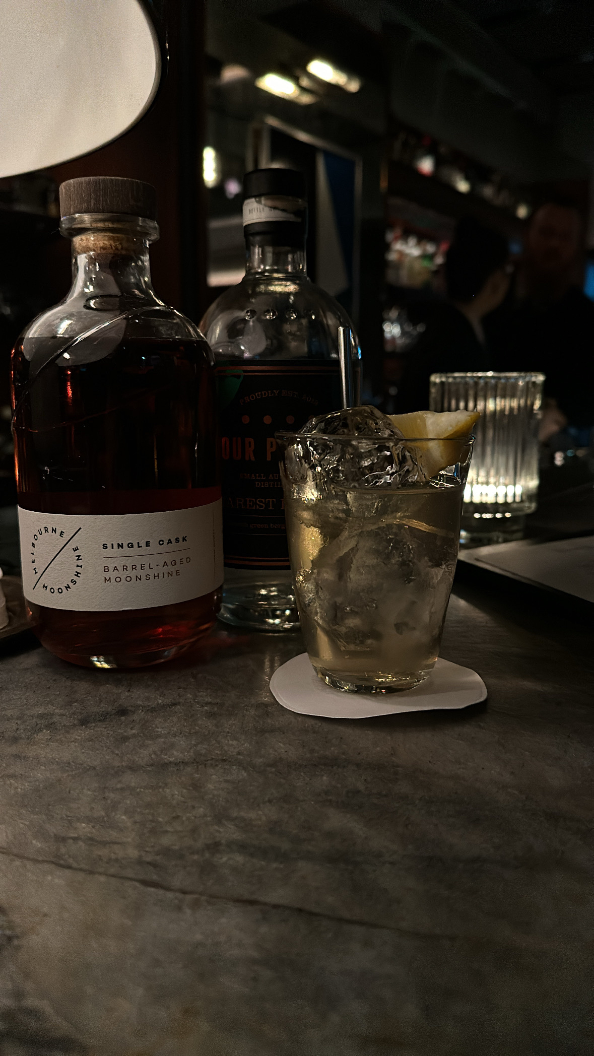 A marble top bar. A glass with a slightly opaque liquid in it is on the bar. A spirit bottle beside it with the label “Melbourne Moonshine. Single cask barrel aged moonshine”. A second spirit bottle is behind the glass and in the background are dimly lit bar shelves.