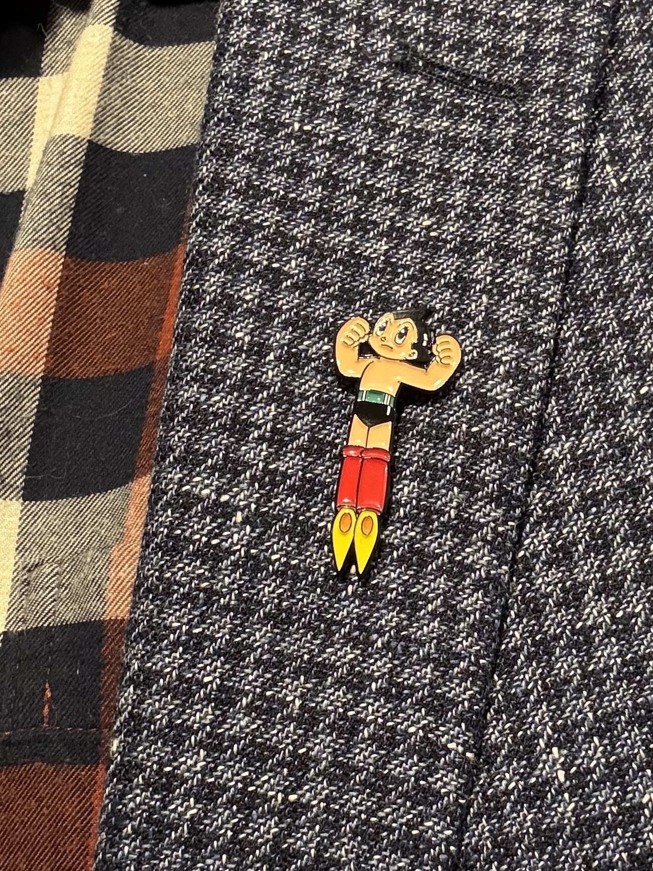 An enamel badge of Astro Boy, the manga character. It’s pinned to a blue check jacket and a dark check shirt is visible under the jacket. 