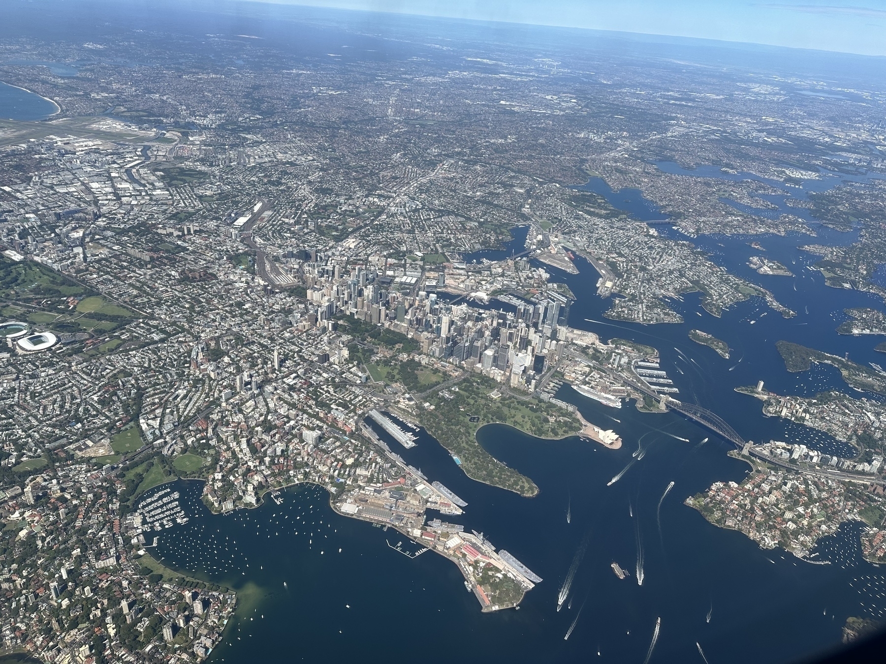 An aerial view of Sydney Harbour. The Sydney opera house, harbour bridge, circular quay, a cruise ship and the domain gardens are all visible as is the CBD and surrounds. The harbour is a deep blue and it’s a clear sunny day.
