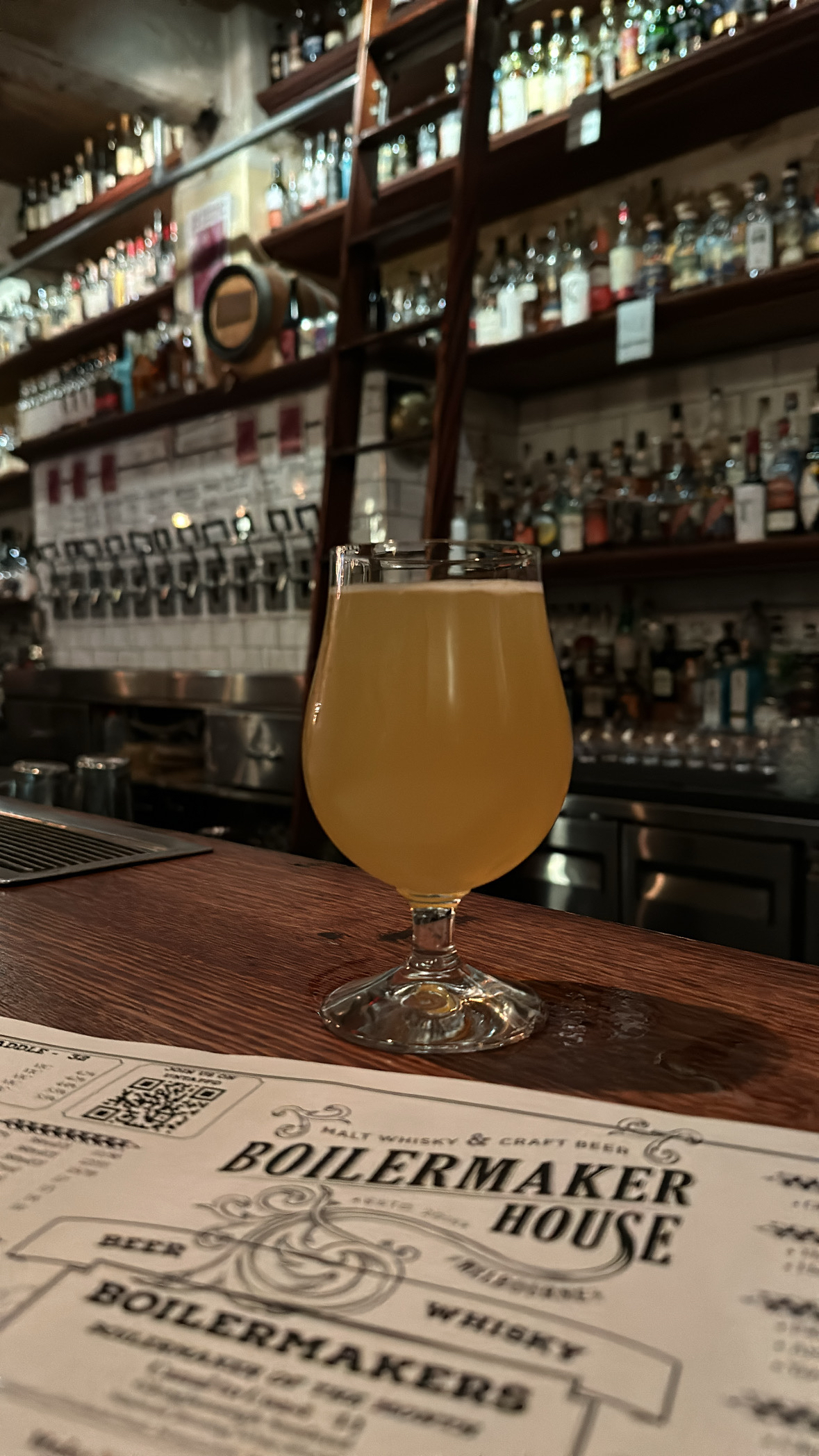 A beer glass sitting on a bar, shelves full of whisky bottles in the background. Part of the menu is in front of the glass displaying the name “Boilermaker House”.