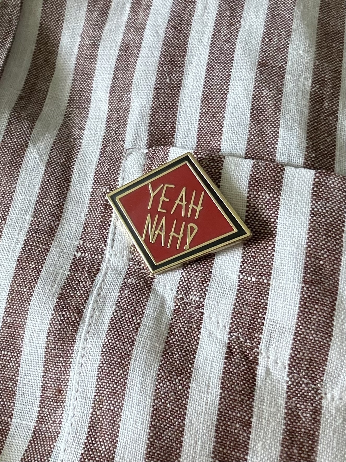 A diamond shaped enamel badge pinned on a brown and white striped shirt. There is a thin black border on the badge, red background, and gold lettering which says “Yeah Nah!”