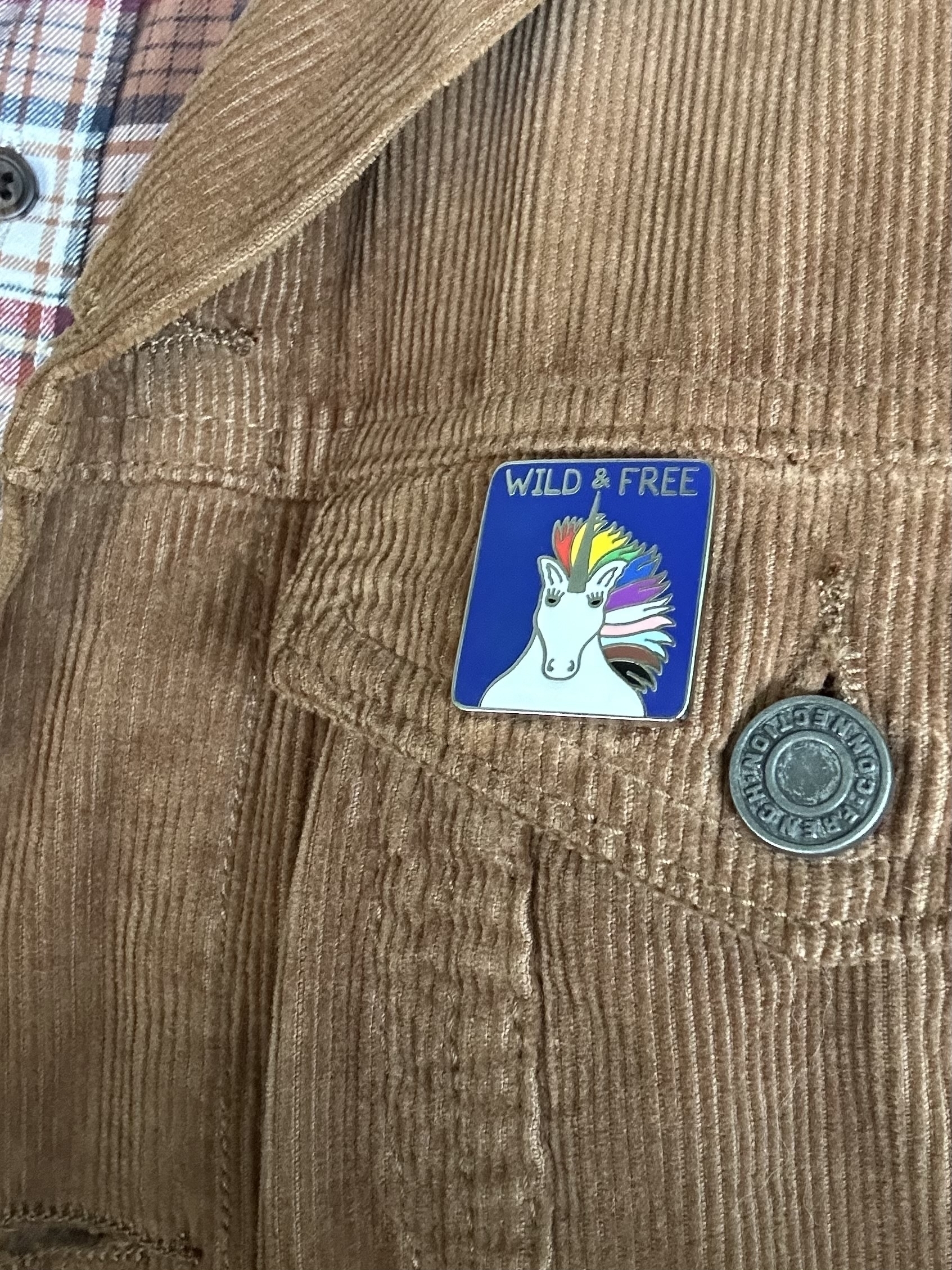 An enamel badge pinned on a brown corduroy jacket. The badge has a blue background and a white unicorn with the LGBTIQ rainbow colours as its mane. The words “Wild & Free” are across the top.