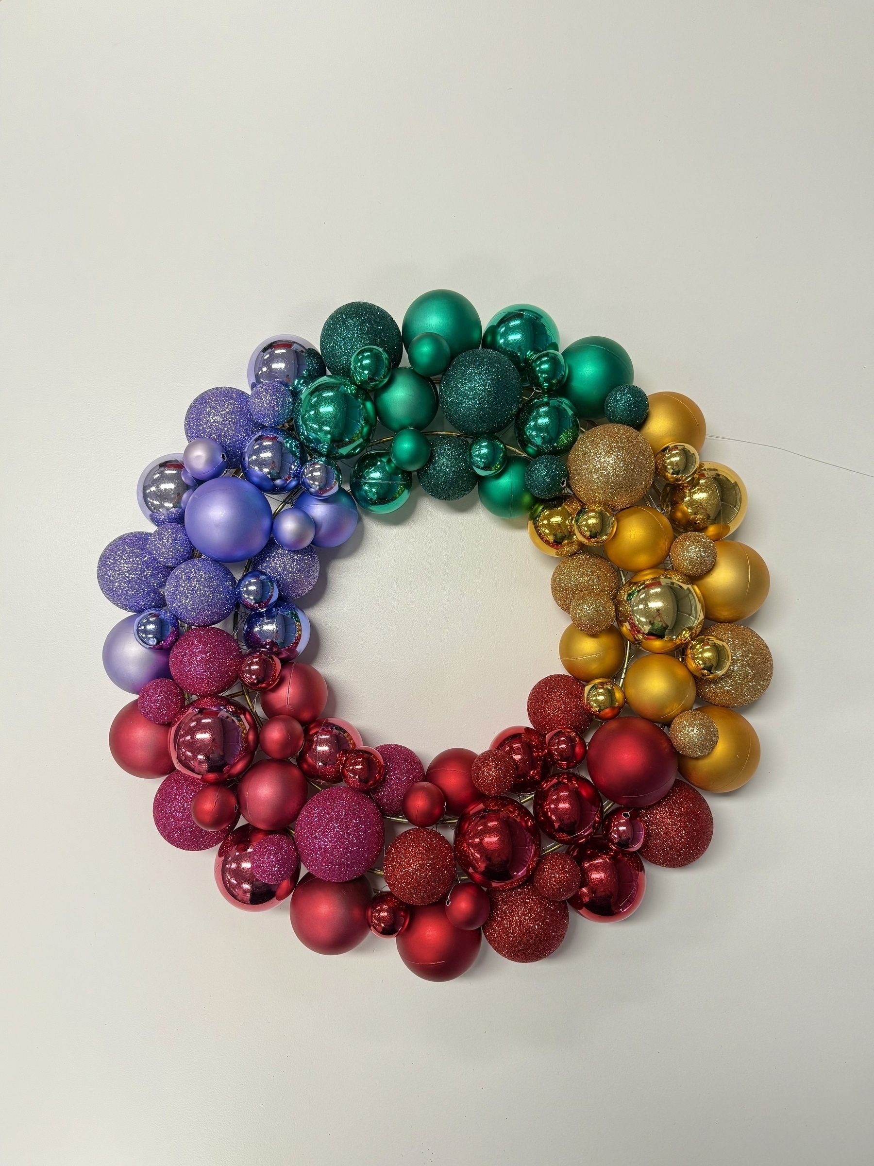 A Christmas wreath made up of different sized spherical baubles, some with glitter, on a round wire frame. The baubles are red, yellow-gold, green, and light purple.