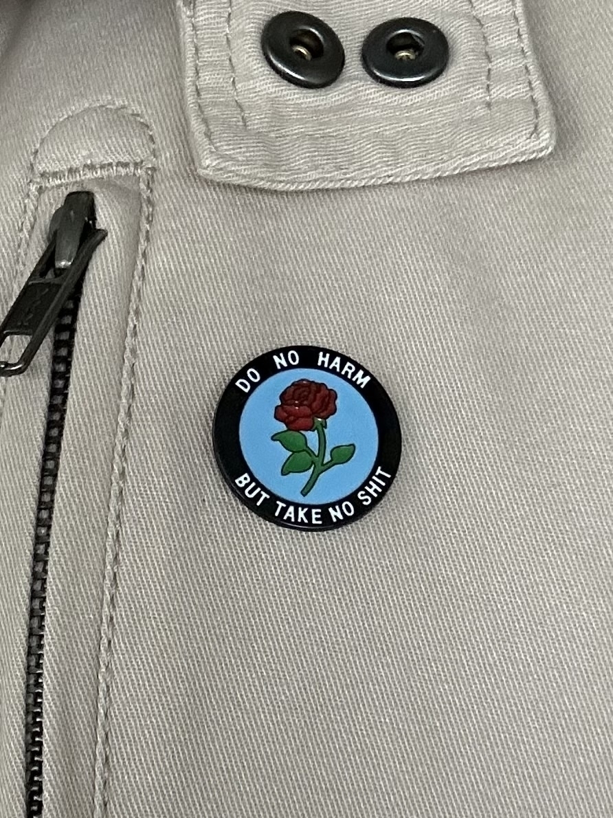 A round enamel badge pinned to a cream jacket. A black ring around the outside with “Do no harm but take no shit” in white letters. Inside the ring is a blue background with a red rose on it.