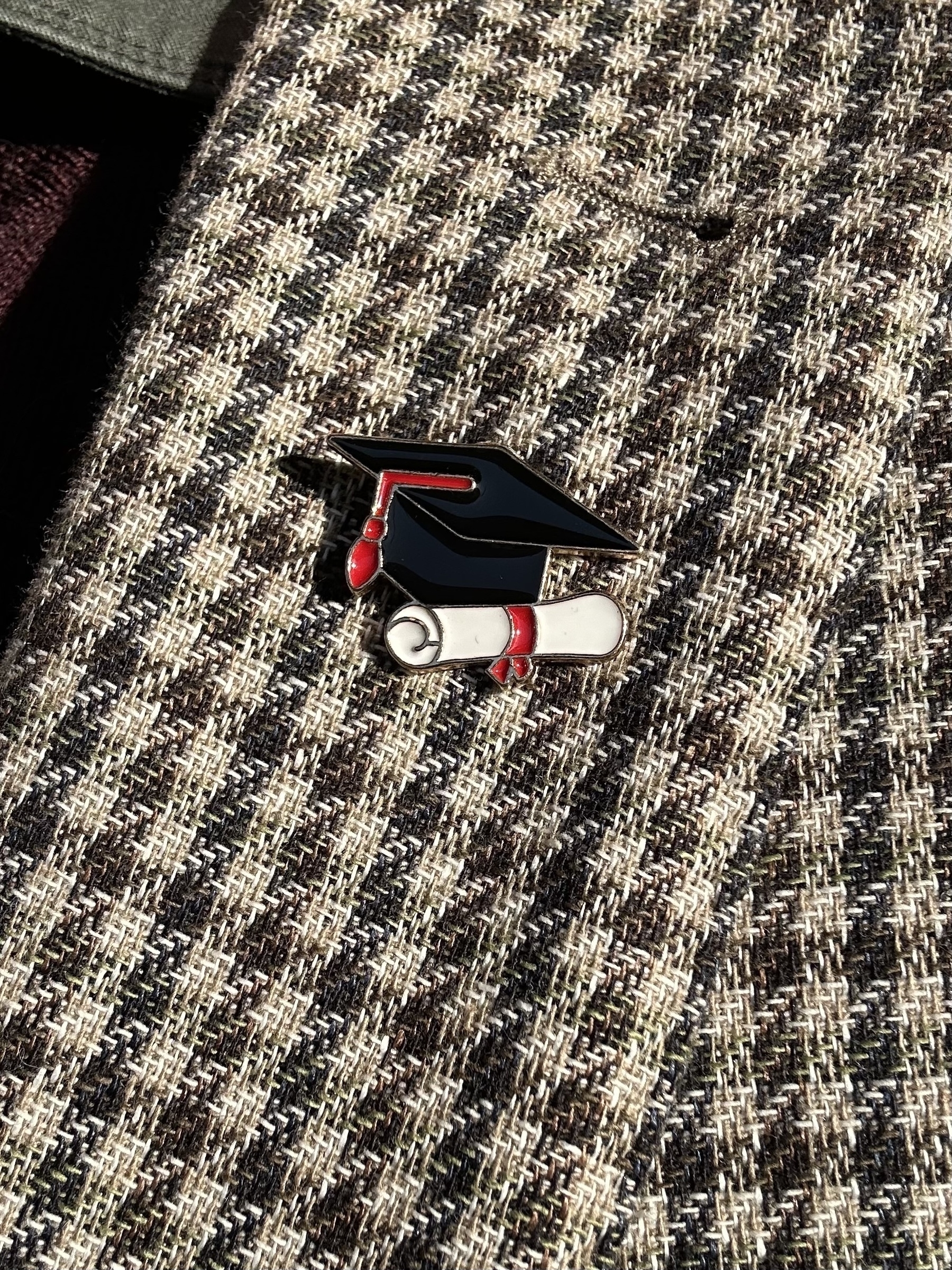 An enamel badge pinned to a green and white check jacket. The badge is a black mortar board with red tassel and a rolled white parchment under.