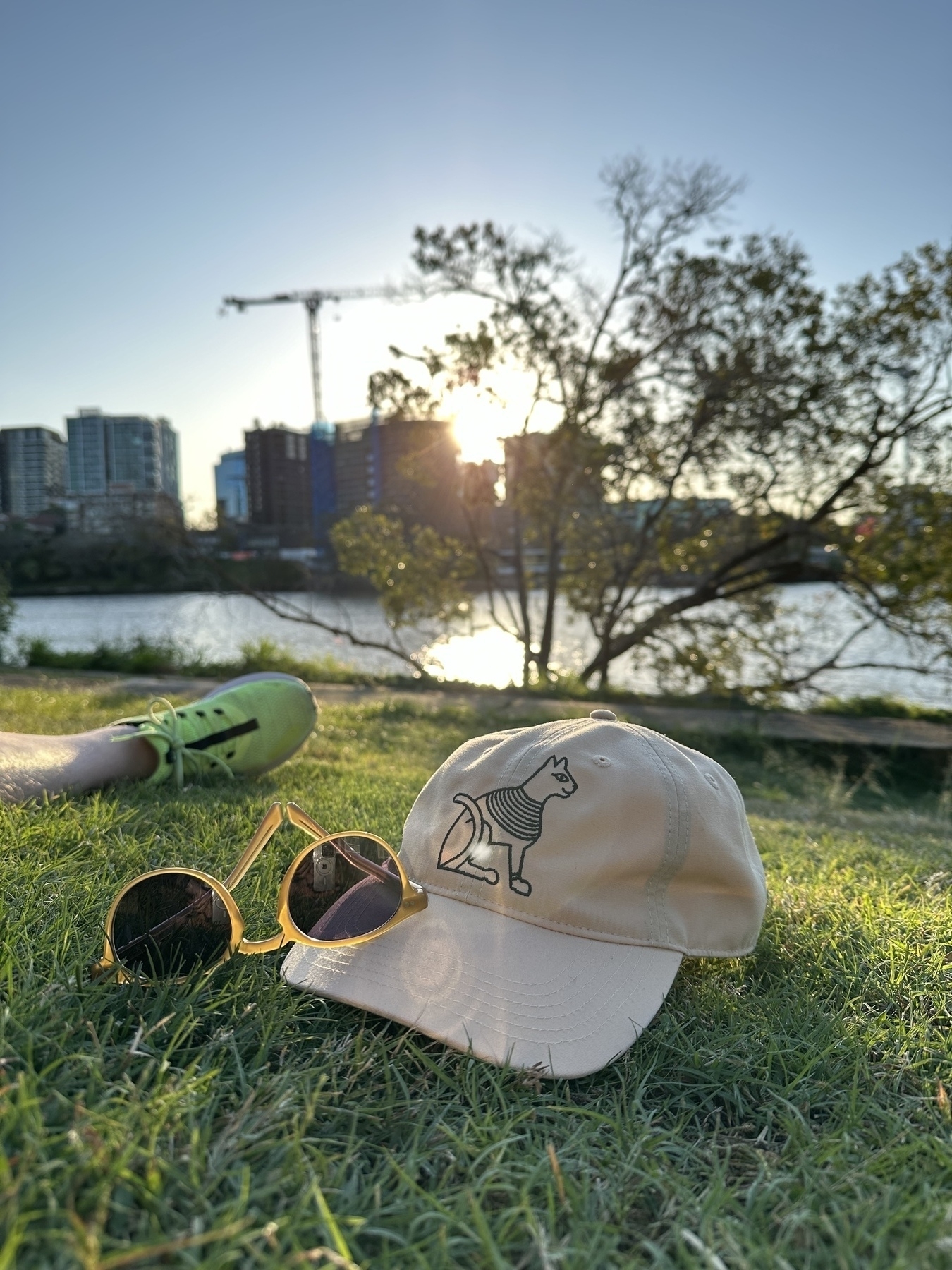 A cap sitting on grass with sunglasses resting on the brim of the cap. In the background is the lower part of a leg wearing a green shoe. Behind that is a tree on the edge of the river and the Brisbane river. On the other side of the river are apartment buildings with a crane over one and the setting sun is visible behind.