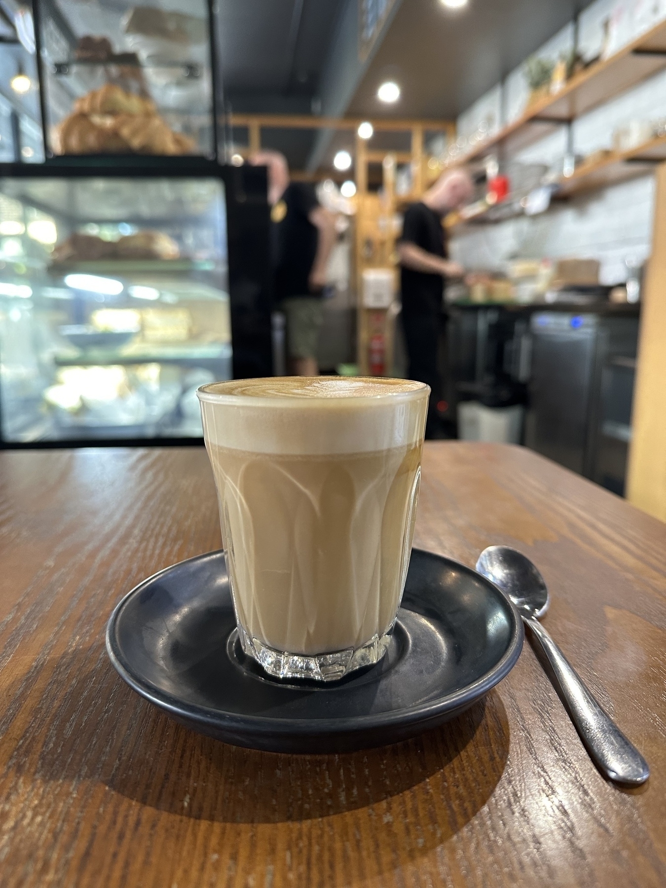 A latte in a glass sitting on a black saucer. A teaspoon is on the wooden table beside the saucer. A blurry background shows two people working behind the cafe counter.