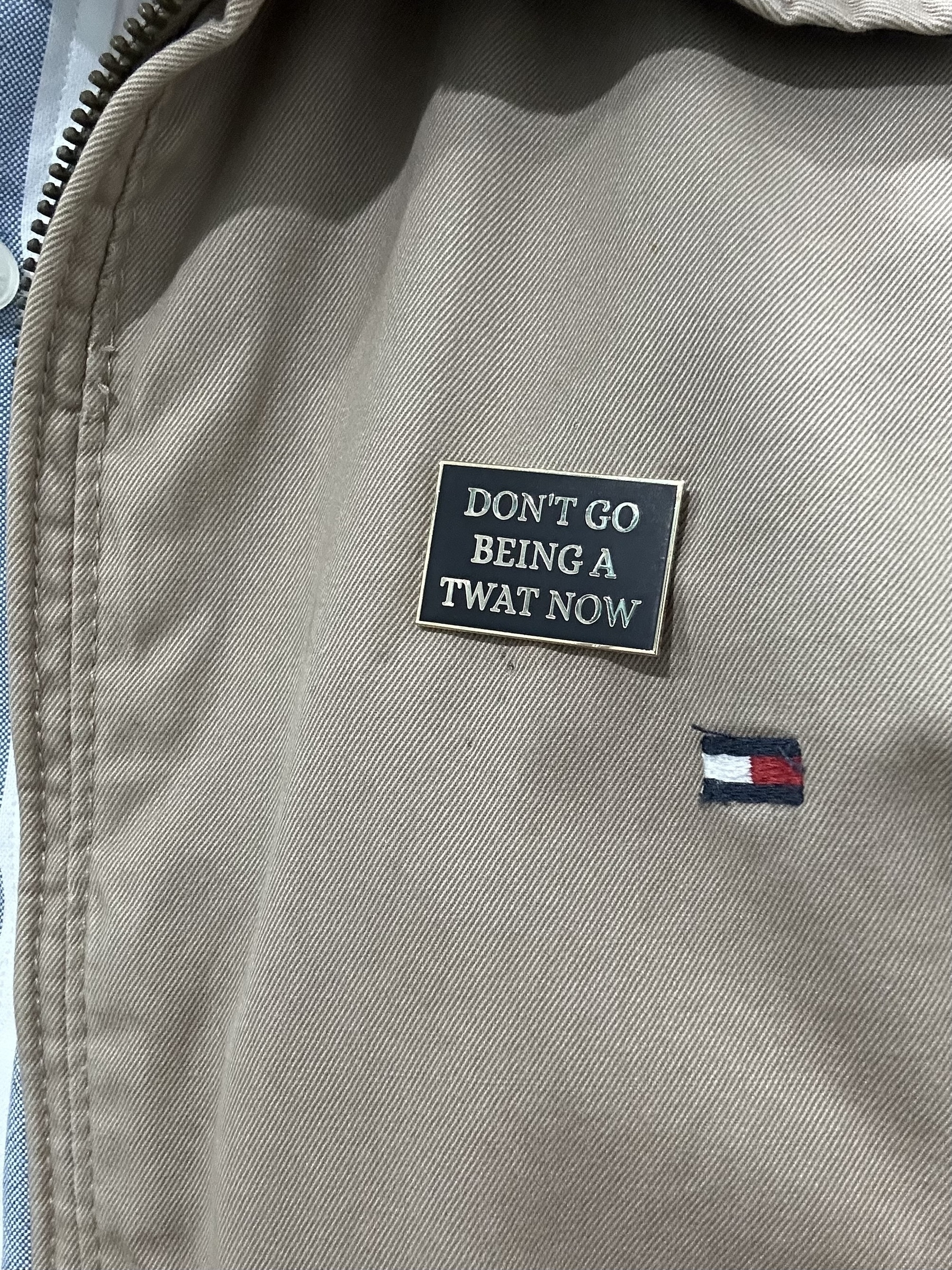 A rectangular enamel badge pinned on a cream jacket. The badge has a black background and gold lettering that says “Don’t go being a twat now”