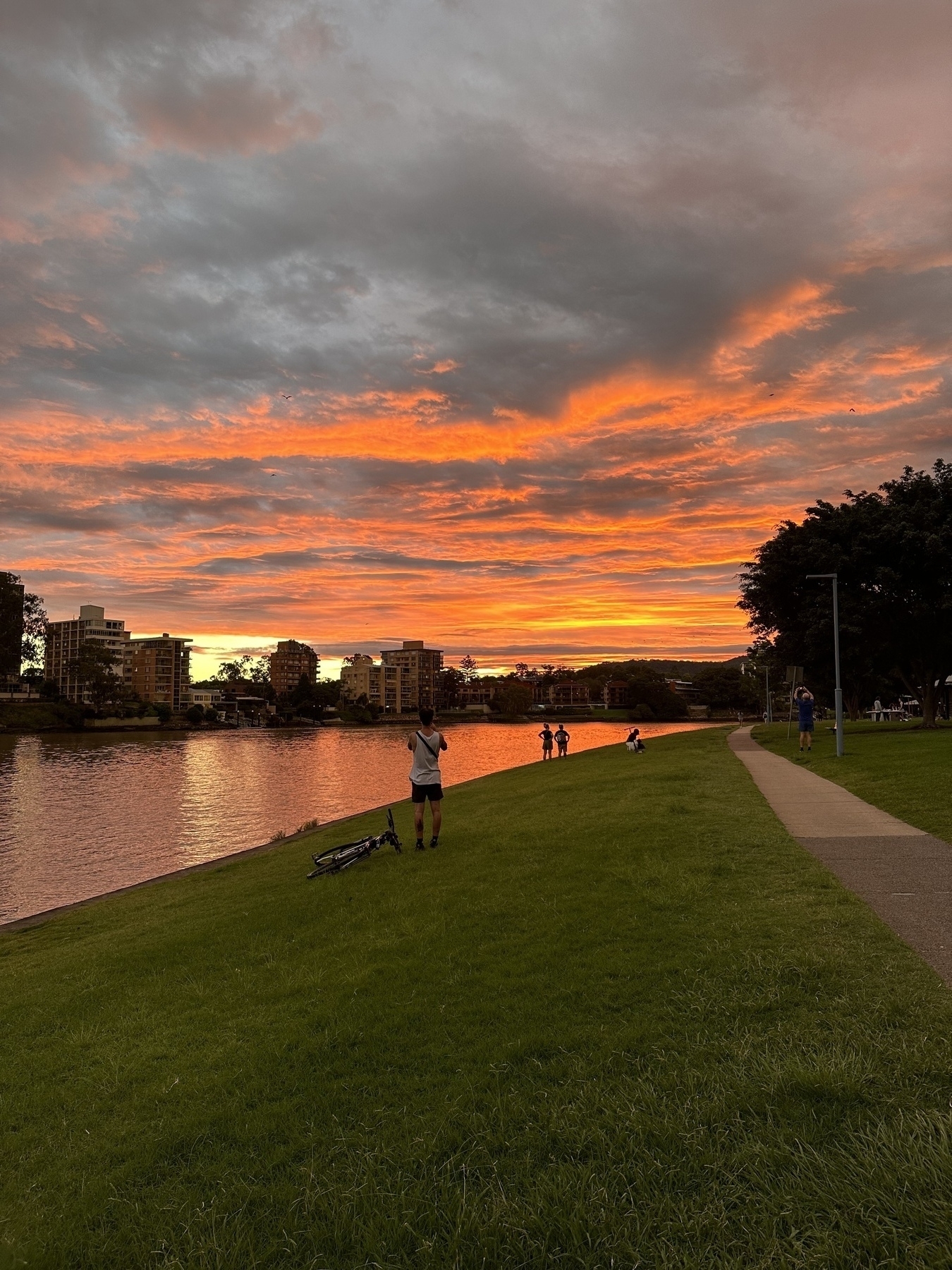 A length of green grass and a cement path in the foreground. A river curving from left to right. On the other side of the river the silhouettes of apartment buildings are visible. Above those is a cloudy sky lit up in a vibrant orange by the setting sun.