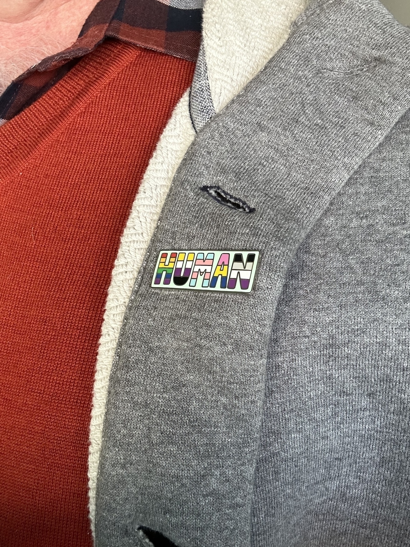 A rectangular enamel badge pinned to a light grey hoodie, part of a burnt orange vest is visible under the hoodie. The badge has HUMAN with the letters some of the various LGBTIQ flag colours.