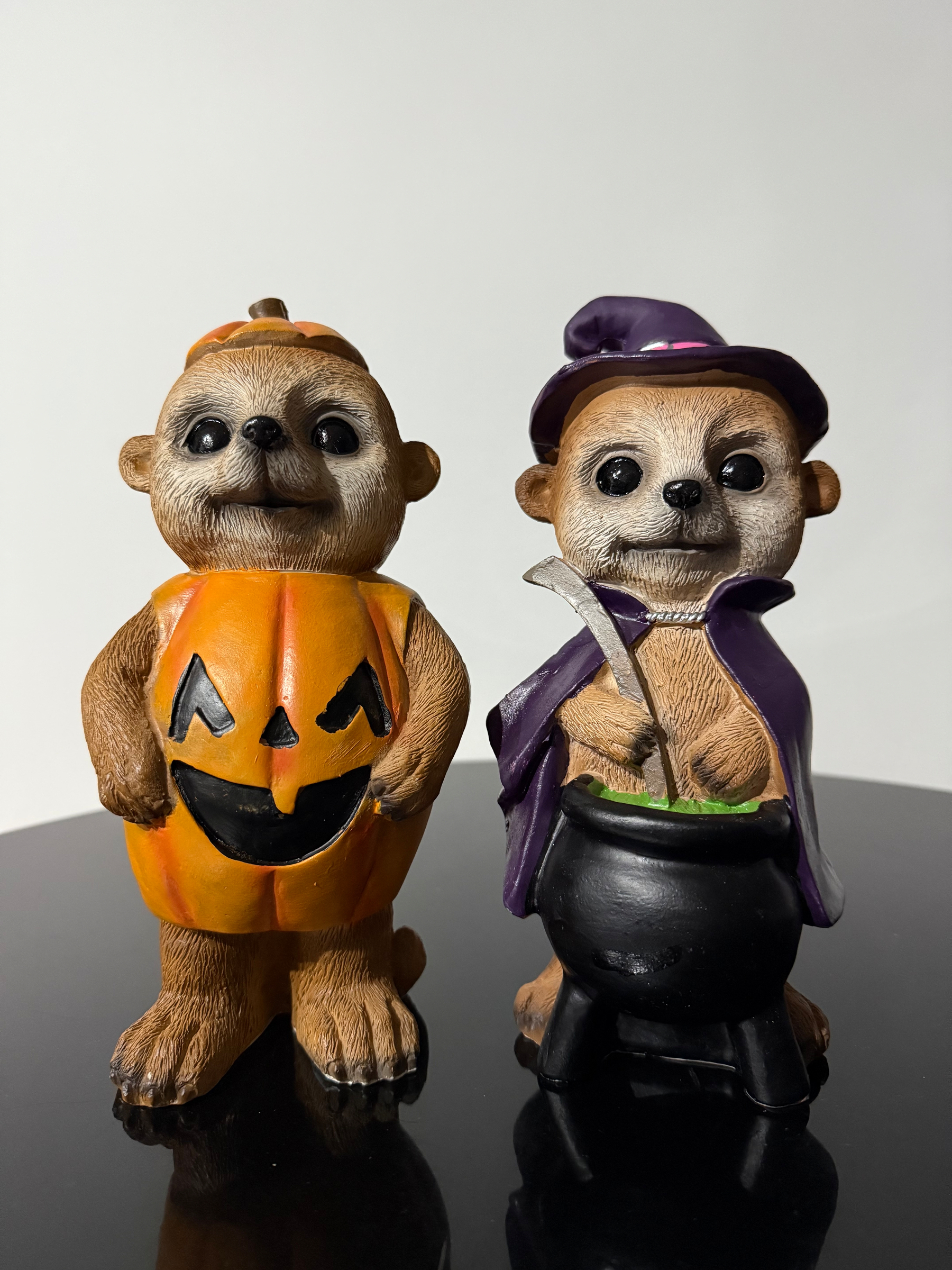 Two plaster models of meerkats, about 20cm in height. The one on the left is wearing a pumpkin like a barrel, with the cut off top as a hat. The one on the right has a purple witches hat and cloak and is standing behind a black cauldron they’re stirring with a big ladle.