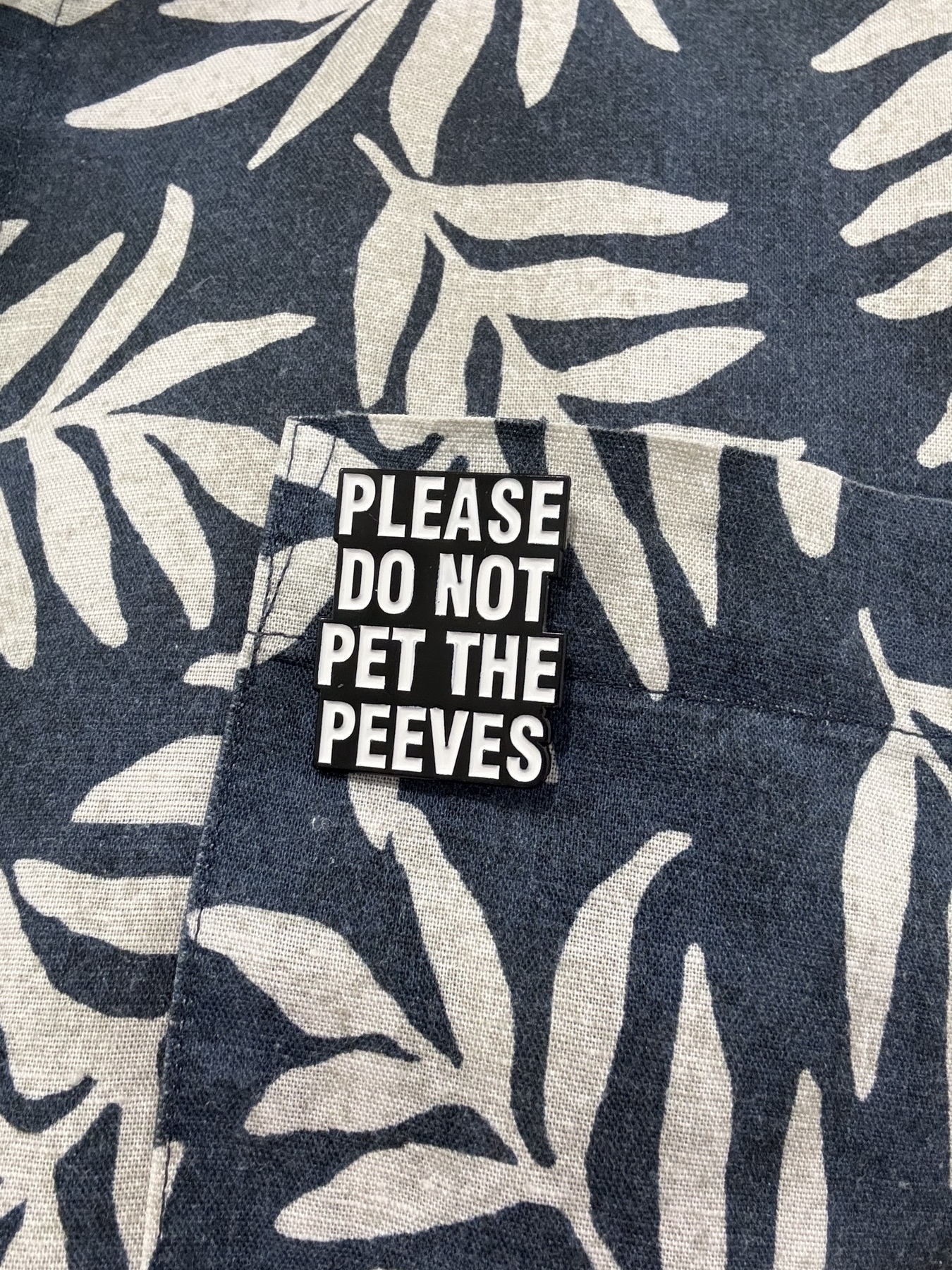 An enamel pin on a blue and white shirt. The pin has a black background with white letters in all caps which say - please do not pet the peeves.