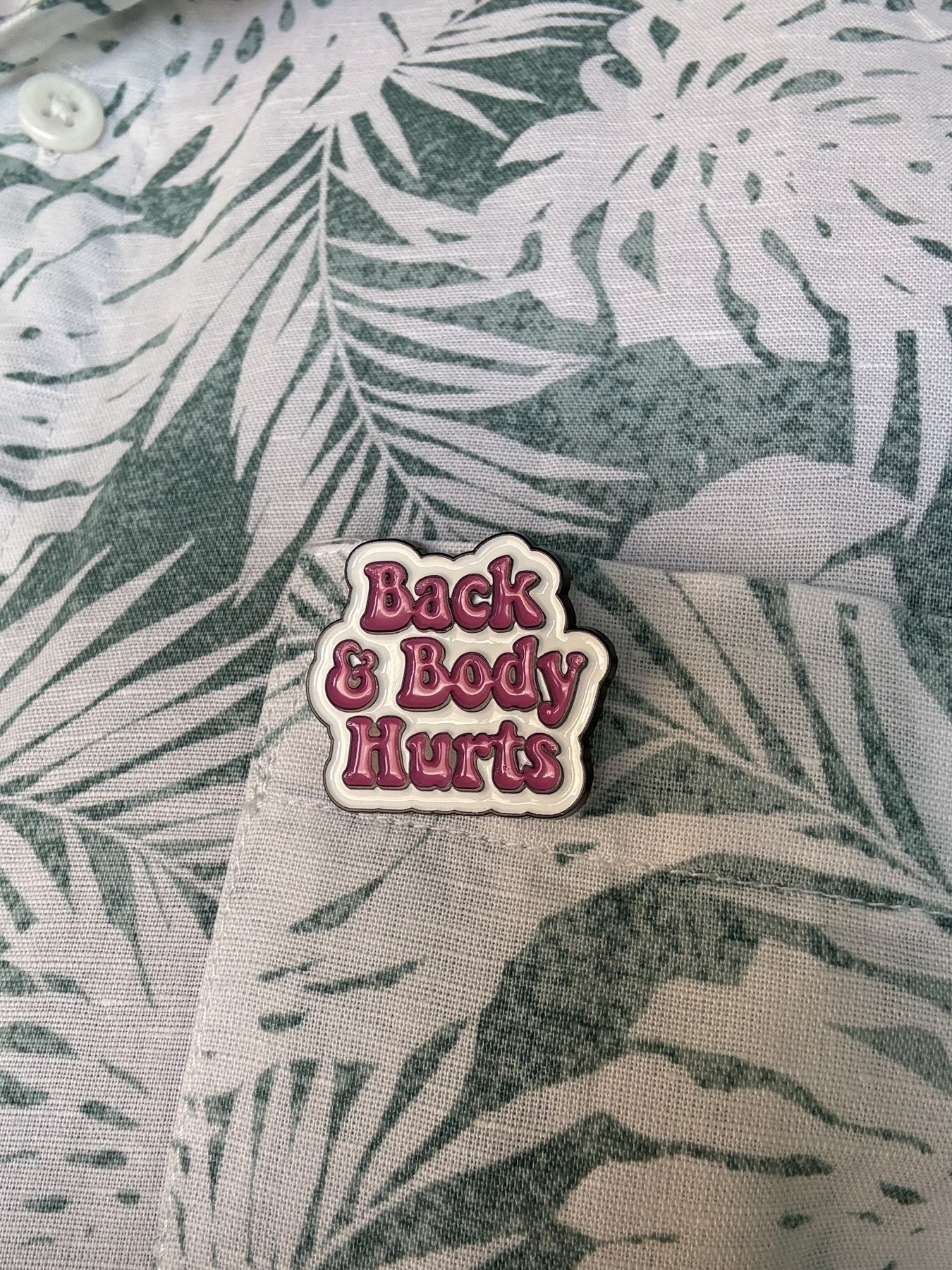 An enamel badge pinned on a green and white Hawaiian print shirt. The badge has a white background and in pink lettering says: Back & Body Hurts