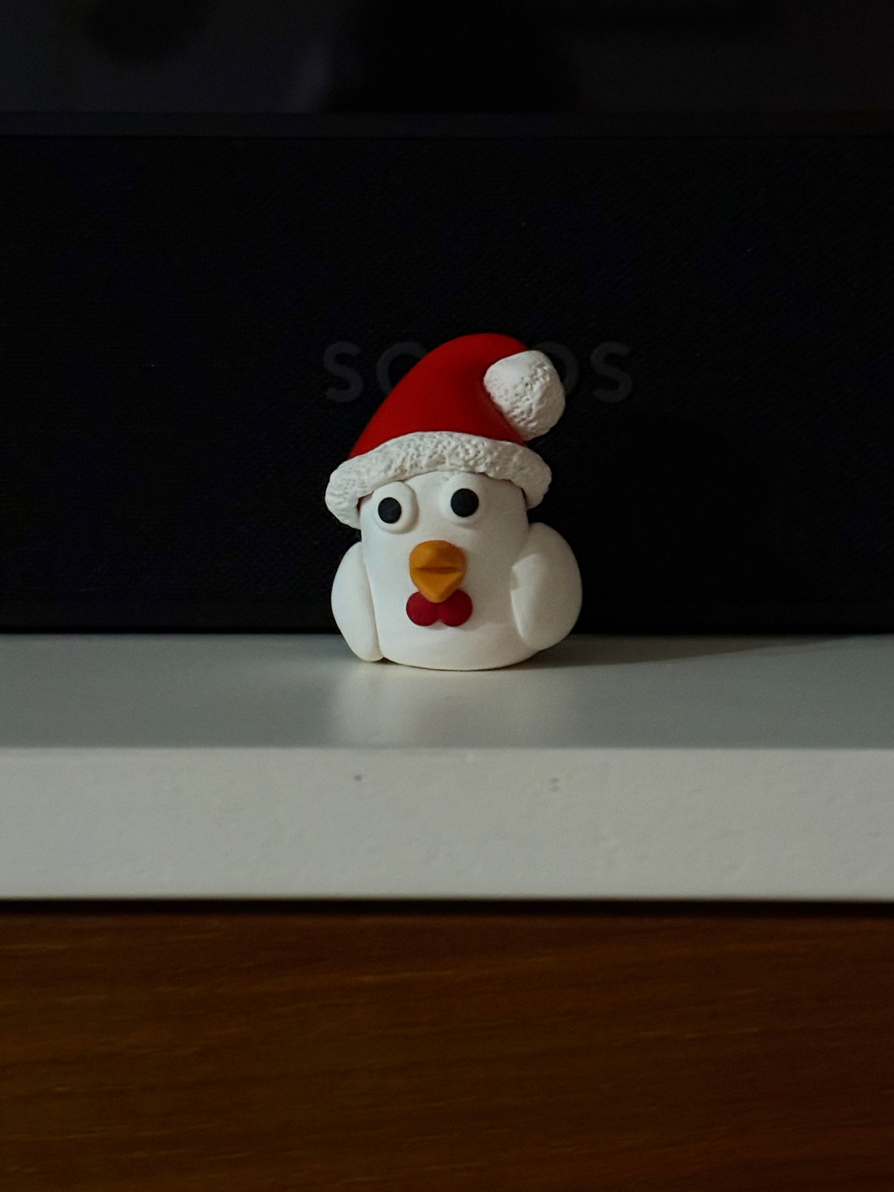 A small white chicken figurine. The chicken looks like it is sitting down as no legs are visible. It has big googly eyes, and is wearing a red bow tie and a Santa hat.&10;&10;It’s sitting on a tv unit with a white top and wood beneath. Behind it is a black Sonos speaker.