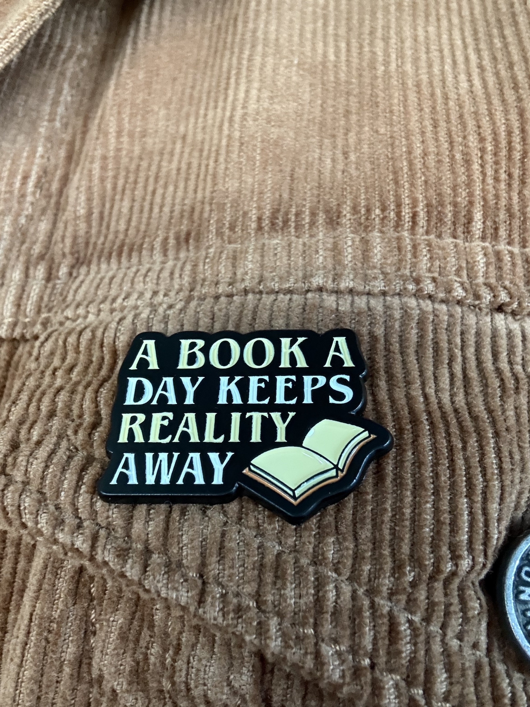 An enamel badge pinned to a brown corduroy jacket. The badge says - A book a day keeps reality away. There is an open book image in the bottom right corner.