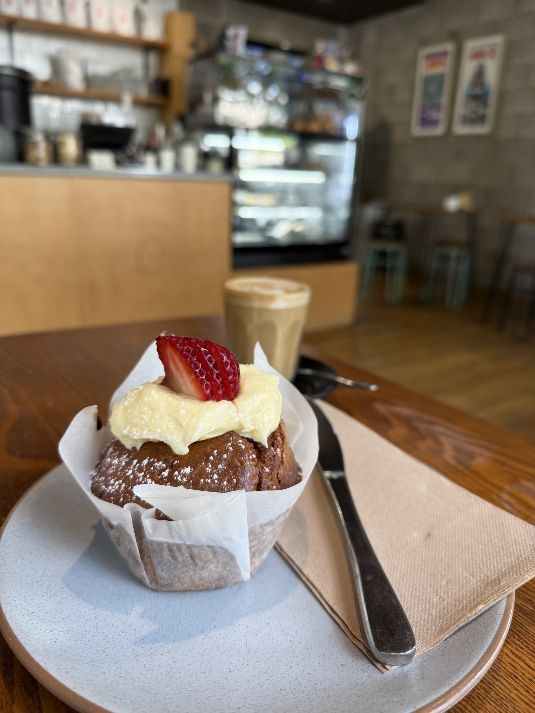 A table in a cafe with a muffin in baking paper wrap in the front of the photo. A large smear of creamy icing with sliced strawberry on top. Behind is a latte in a glass. In the background is the counter and cabinet with treats.