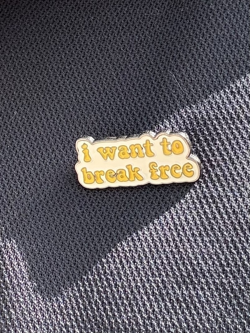 An enamel badge with yellow writing on a white background. It says - i want to break free