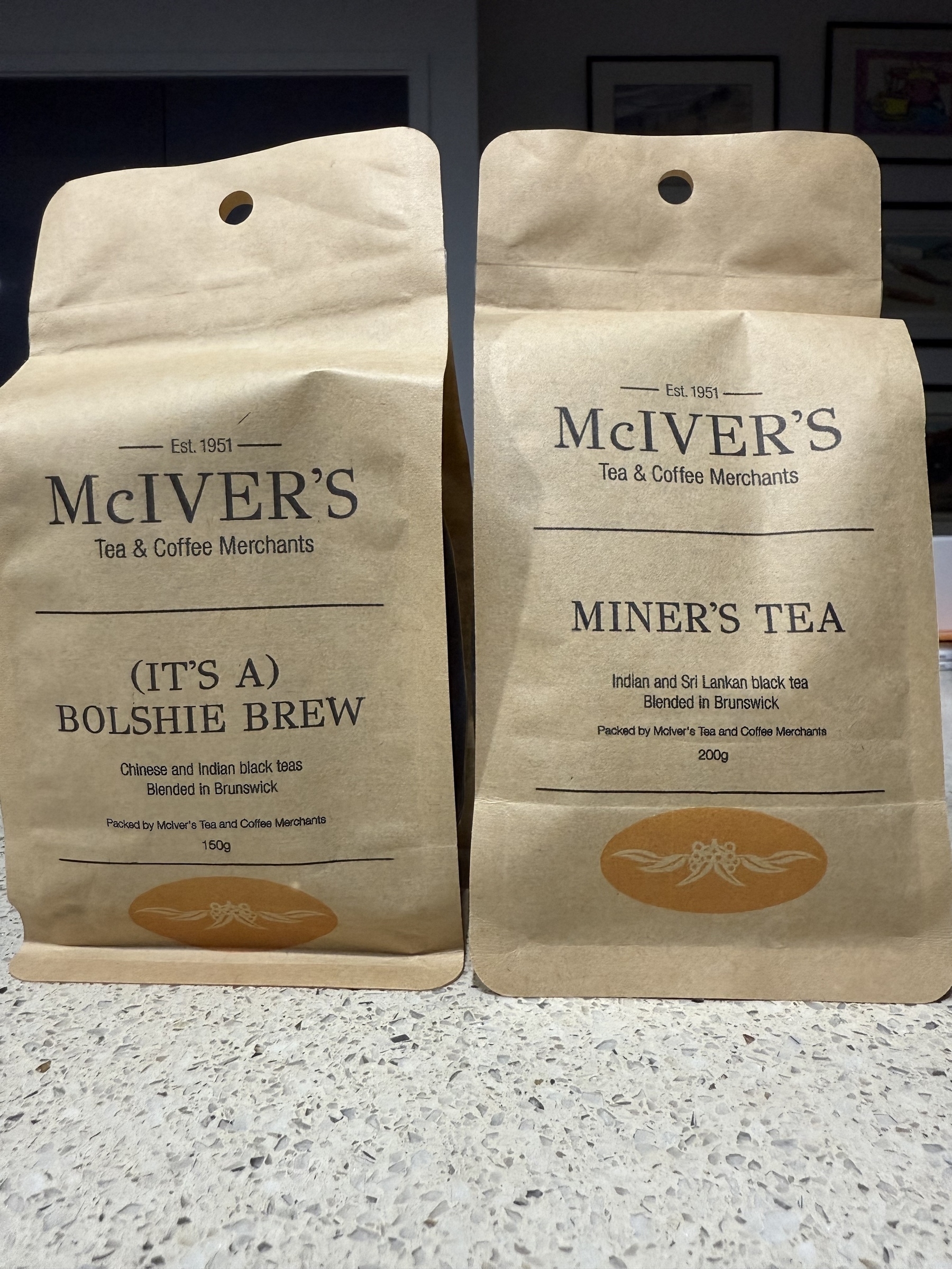 Two brown paper packages with tea inside. The labelling is the same for both and the top has two lines - “McIVER’S” and “Tea & Coffee Merchants” underneath. The bottom half has the name of the tea and indicates the tea blend and where it was blended. The packet on the left is “(It’s a) Bolshie Brew” and on the right is “Miner’s Tea”.