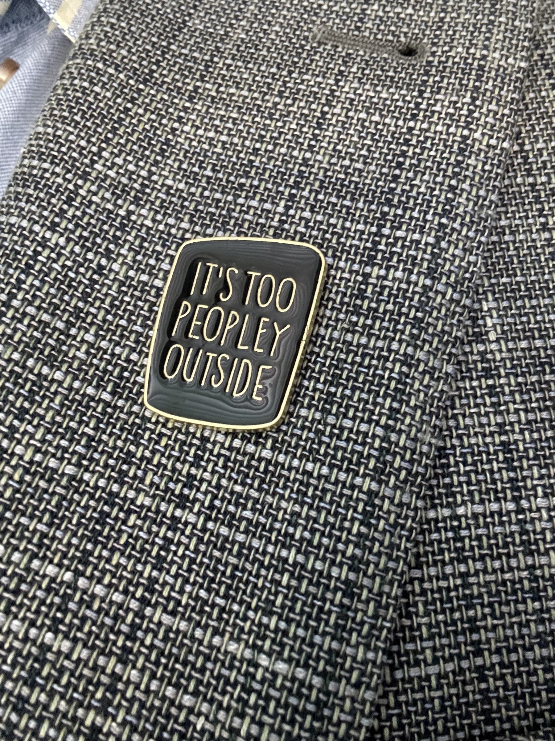 An enamel badge pinned on a jacket. The badge has a black background and in all caps gold letters says - It’s too peopley outside.