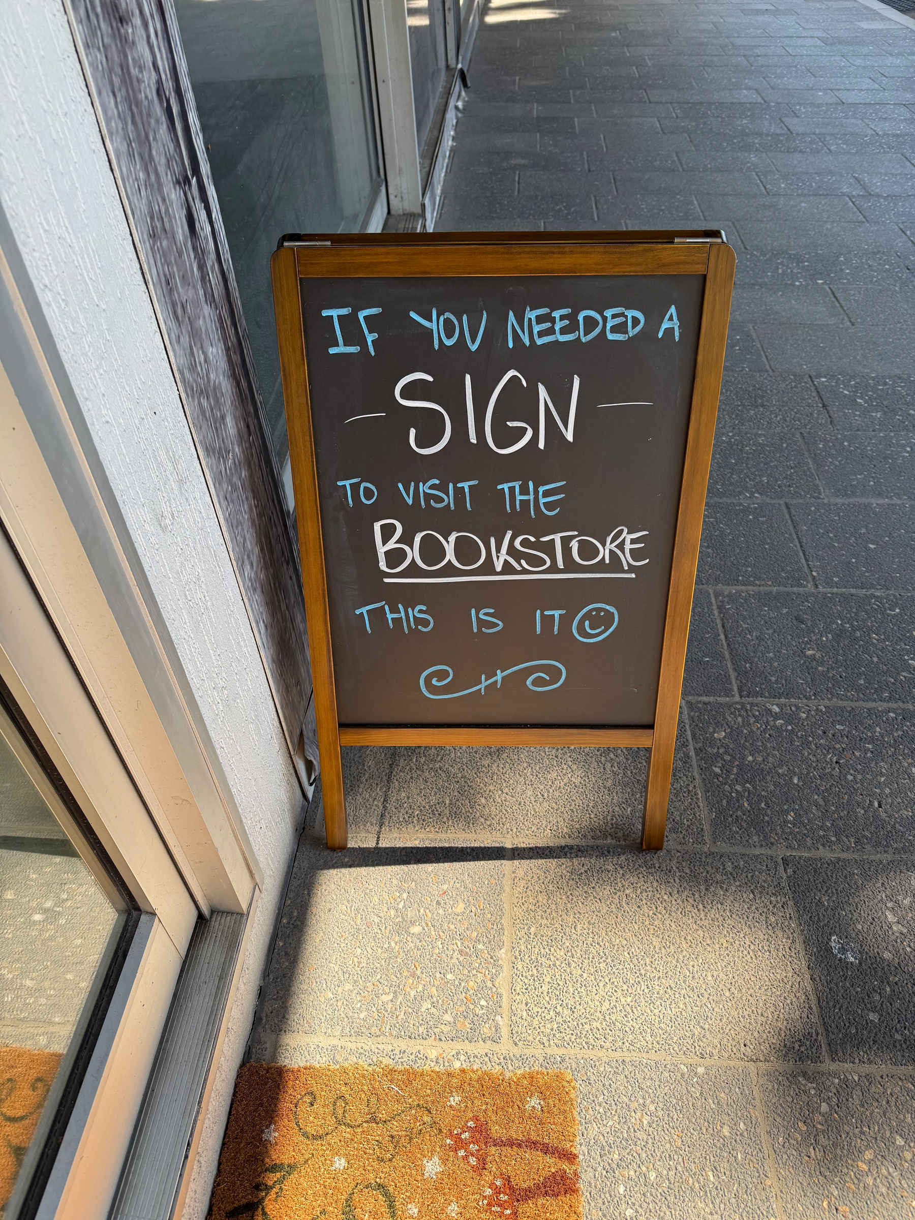 A chalk noticeboard on a footpath outside a shop door. The board says - If you need a sign to visit the bookstore this is it.