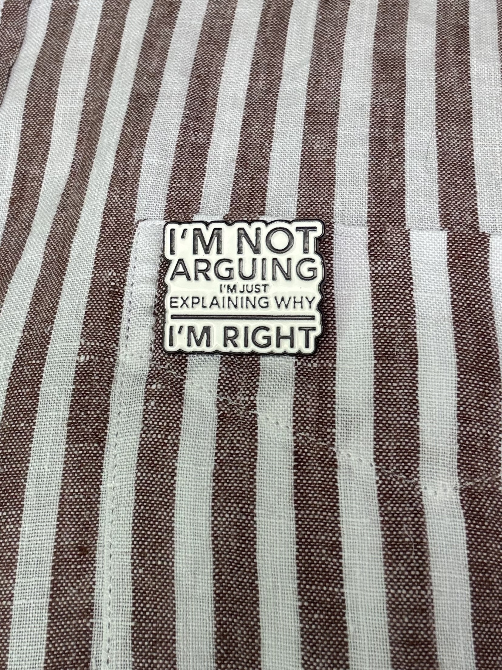 A black and white enamel badge pinned on a brown and white striped shirt. Badge says: I’m not arguing I’m just explaining why I’m right