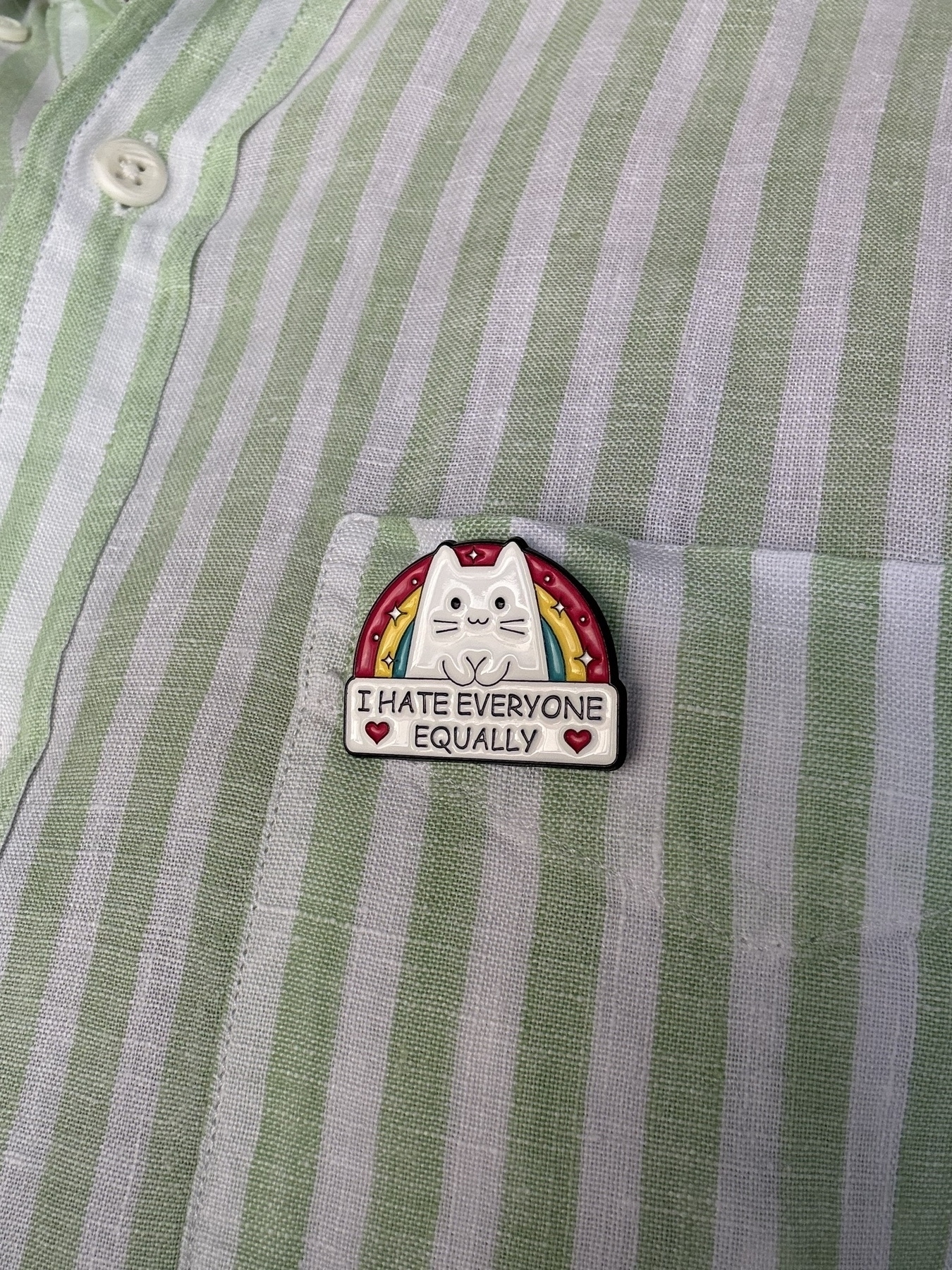 An enamel badge pinned to a green and white striped shirt. The badge has a white cat head with a semi circle green, yellow, red rainbow behind it. The cat’s little paws resting on a rectangular section with the words: I Hate Everyone Equally ❤️