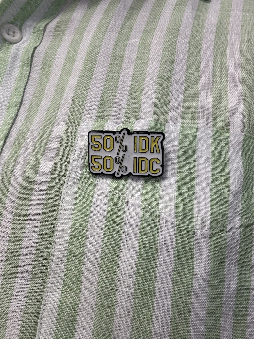 An enamel badge with two lines “50% IDK, 50% IDC” pinned to a green and white striped shirt.