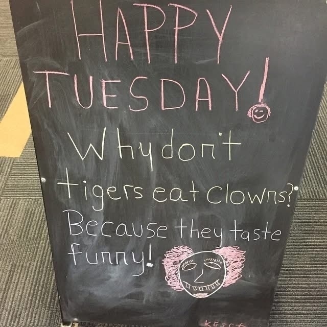 A small noticeboard in a foyer. It says “Happy Tuesday! Why don’t tigers eat clowns? Because they taste funny!” There’s a hand drawn clown at the bottom.