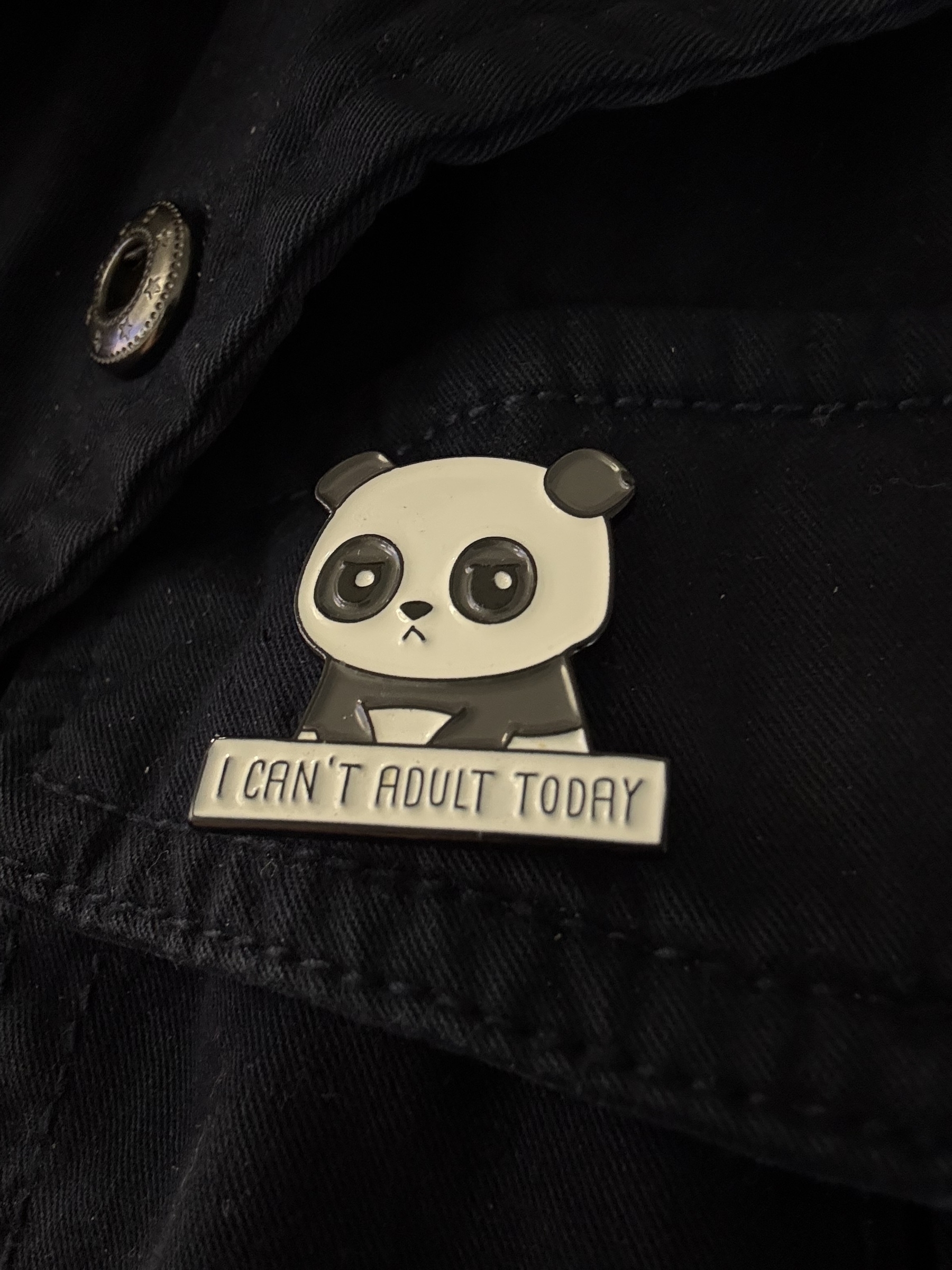 A black and white enamel badge pinned to a dark jacket. The badge has a sad panda sitting down and a rectangular box below with the words - I can’t adult today
