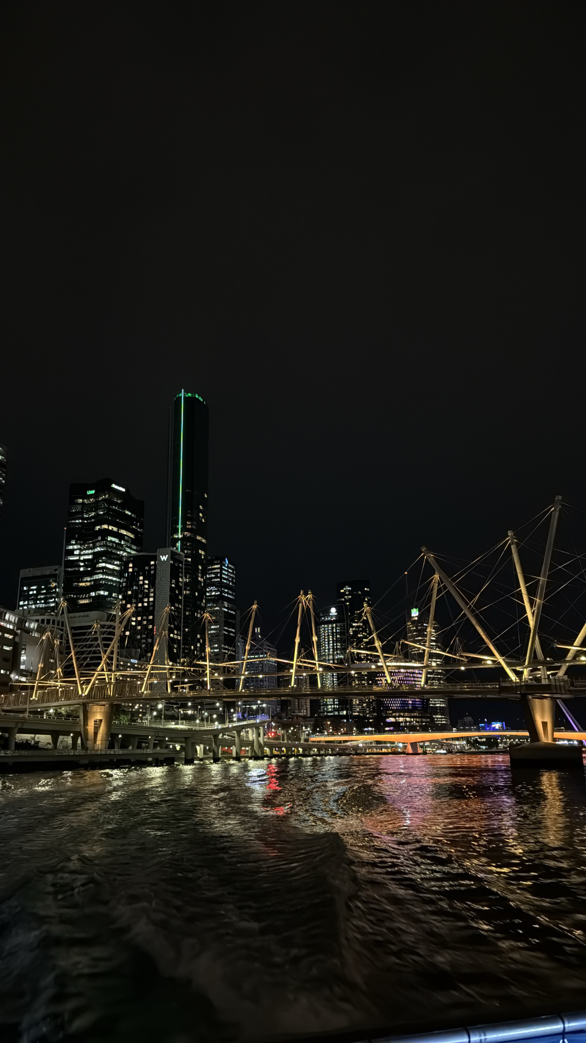 A view from the back of a river ferry at night. The wake visible in the reflected lights of the city. A lit bridge across the river is on the middle with illuminated city buildings behind.