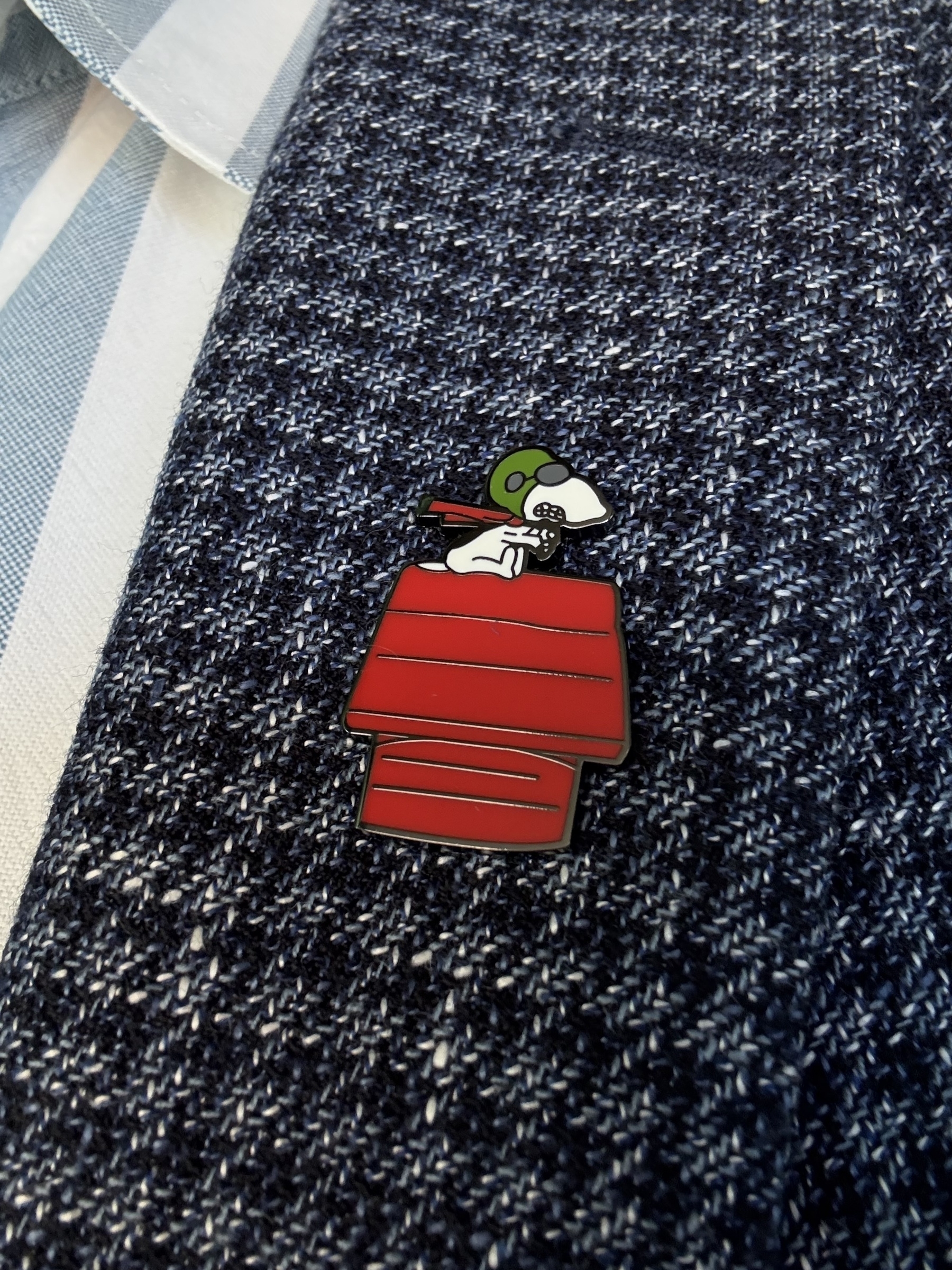 An enamel badge on a blue check jacket. The badge is snoopy as the WWI flying ace sitting/flying his red kennel.