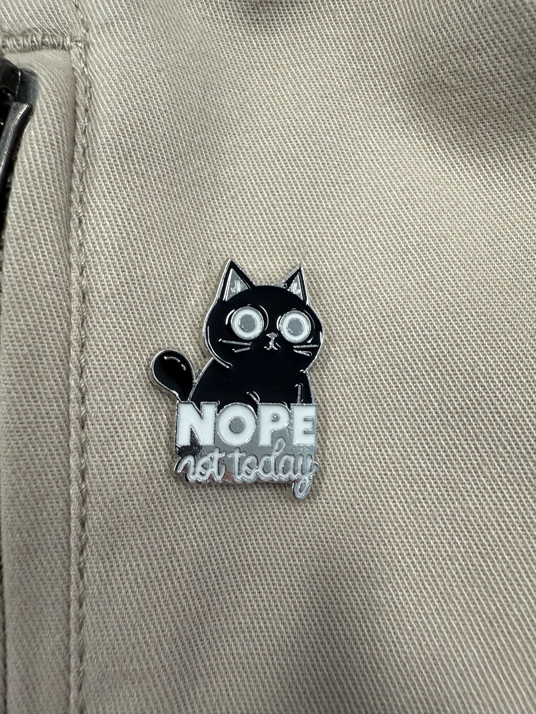 An enamel badge pinned on a cream coloured jacket. The badge is a black cat with big open eyes sitting behind the words - NOPE not today