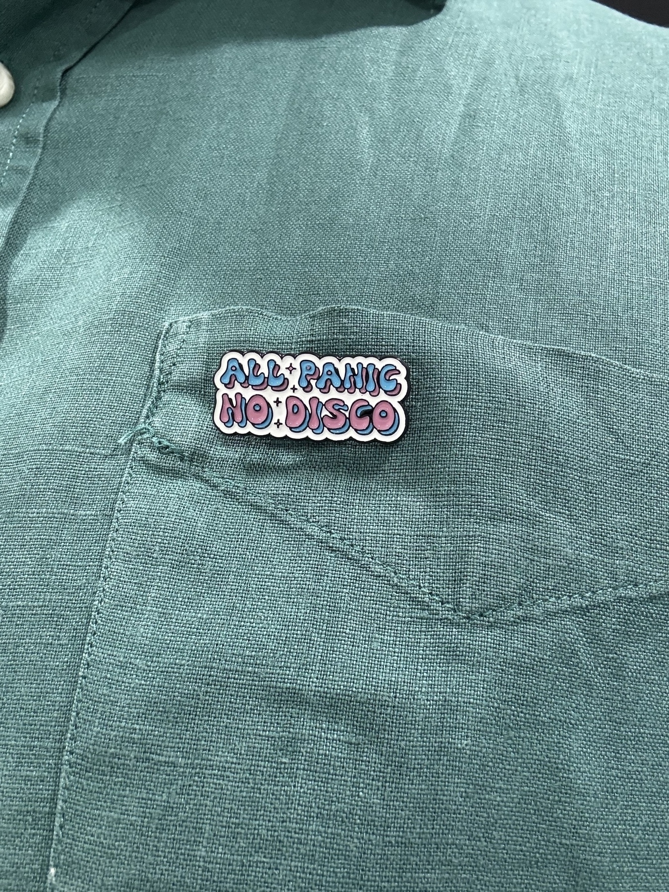 A green shirt with an enamel badge pinned to it. The badge has a white background with two lines of text, top line is blue, bottom is pink. It says: All Panic No Disco