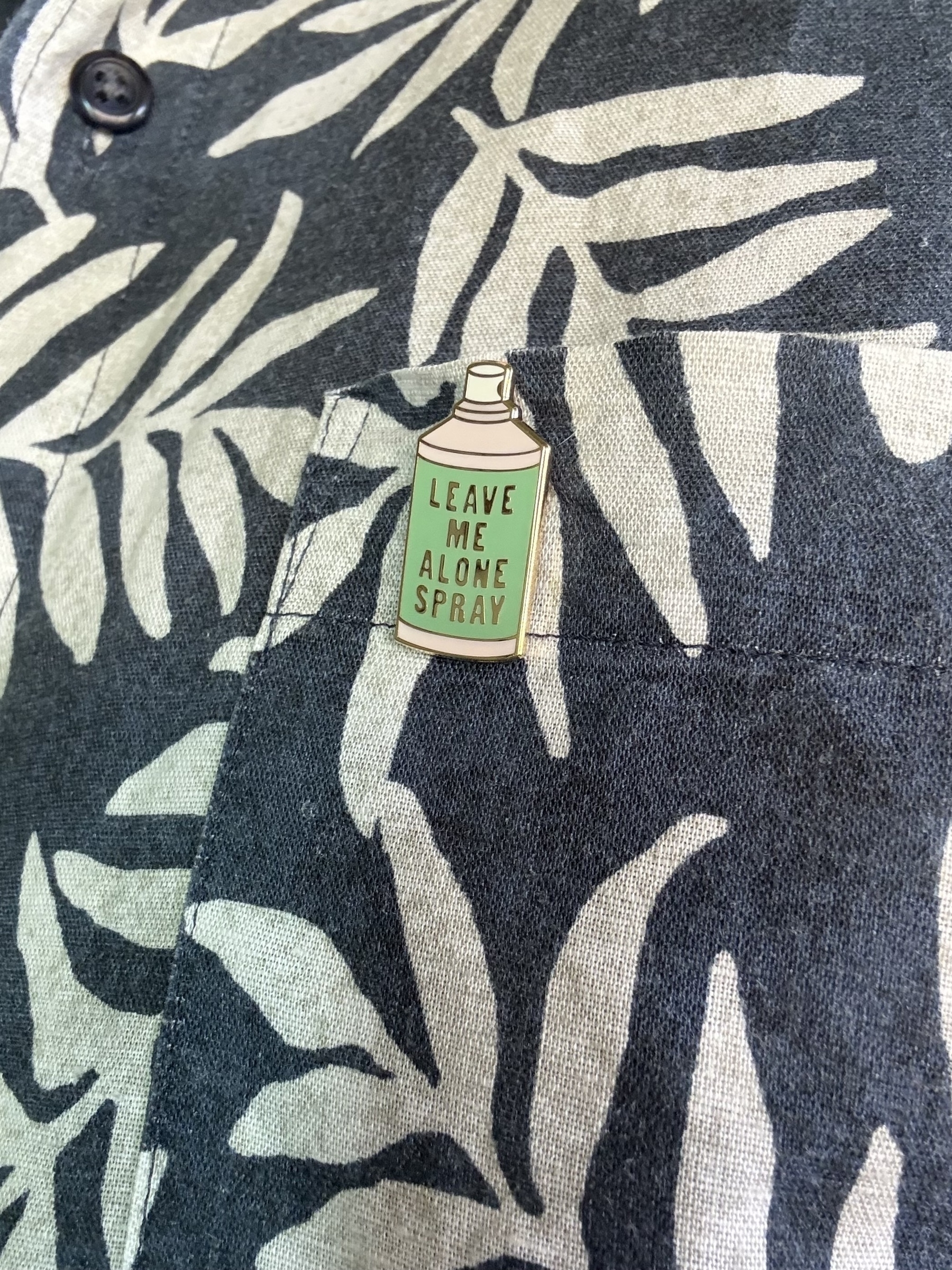 An enamel badge pinned on a blue and white shirt. The badge is green and shaped liked a spray can with the words: Leave me alone spray