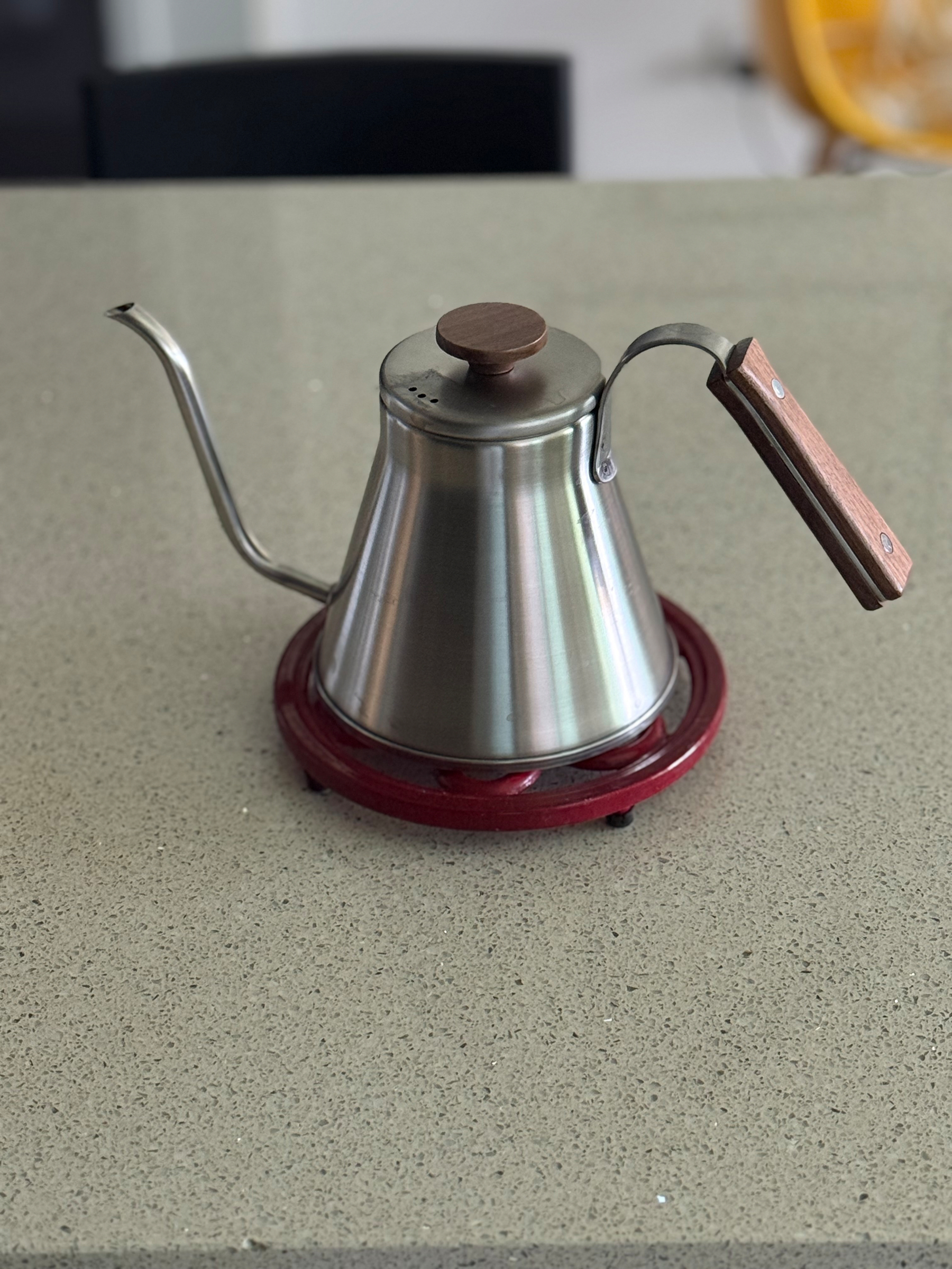 A silvery metal kettle sitting on a red trivet on top of a kitchen bench. The kettle is shaped sort of like an inverted cone and has a long thin curved spout from the bottom. The handle is wood as is the round handle on the lid.