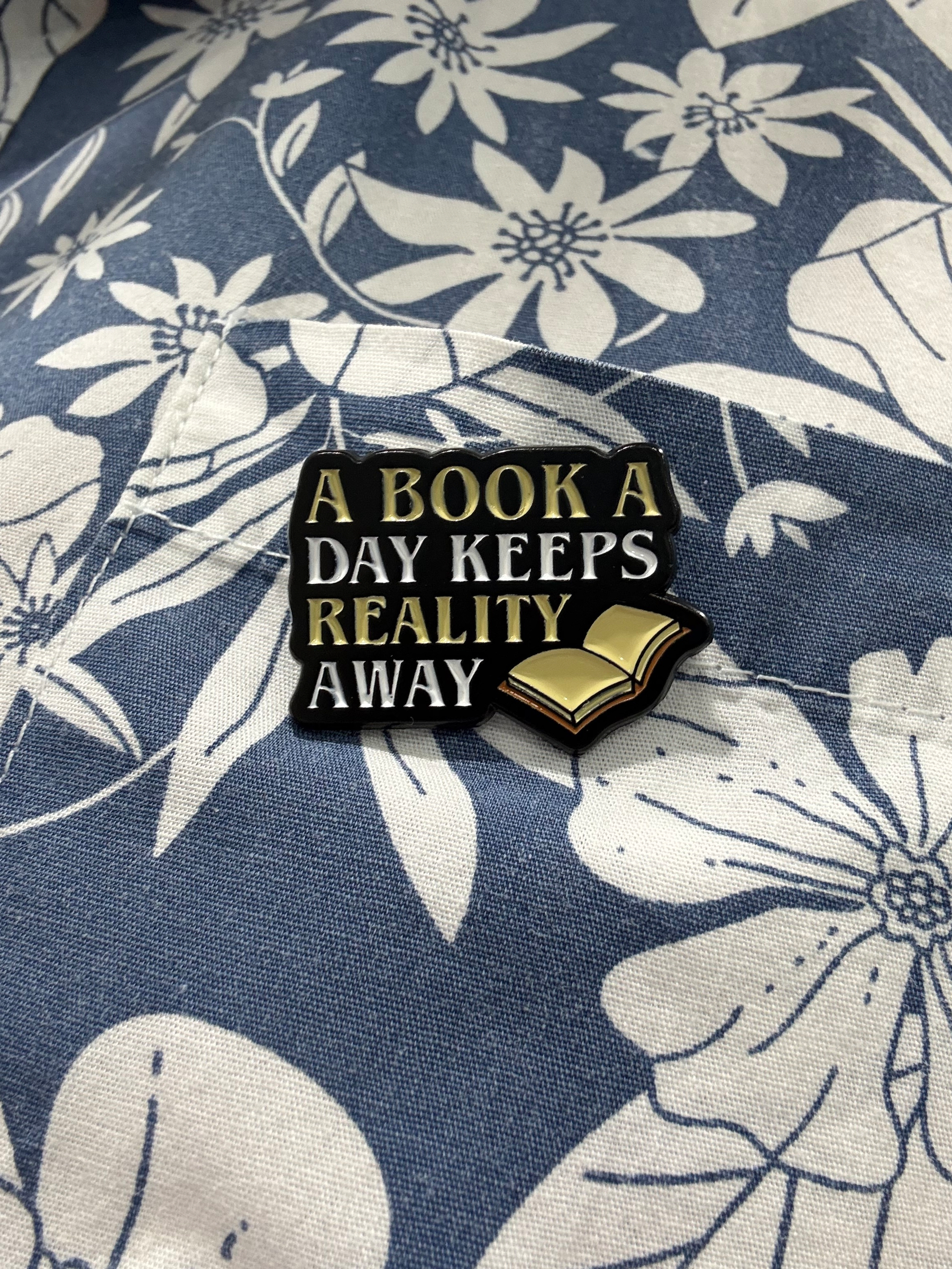 An enamel badge pinned on a blue and white shirt. The badge has the image of an open book in the bottom right corner and says - A book a day keeps reality away.