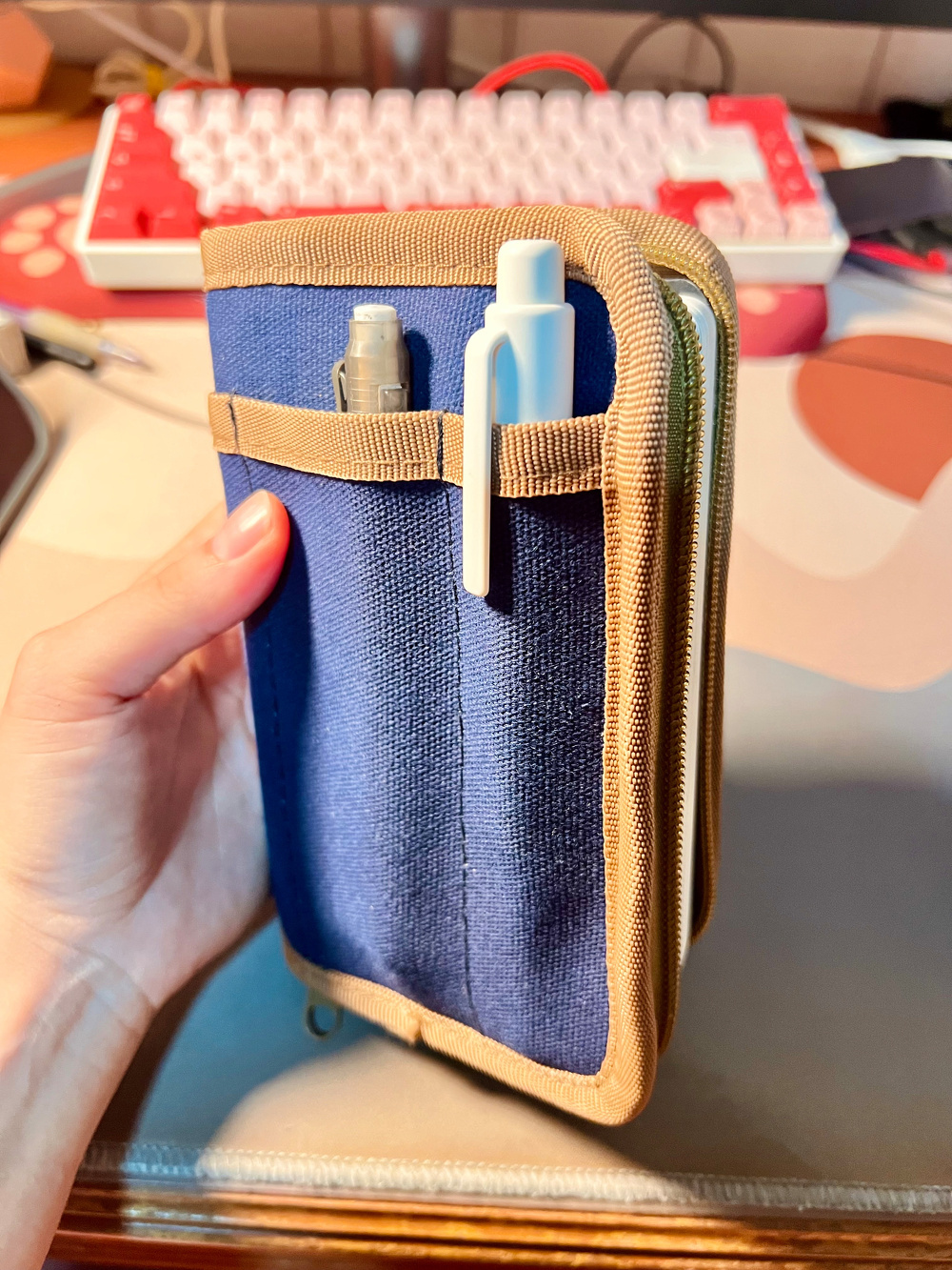 A blue and beige zippered notebook cover with a pencil and a retractable eraser in its front pocket.